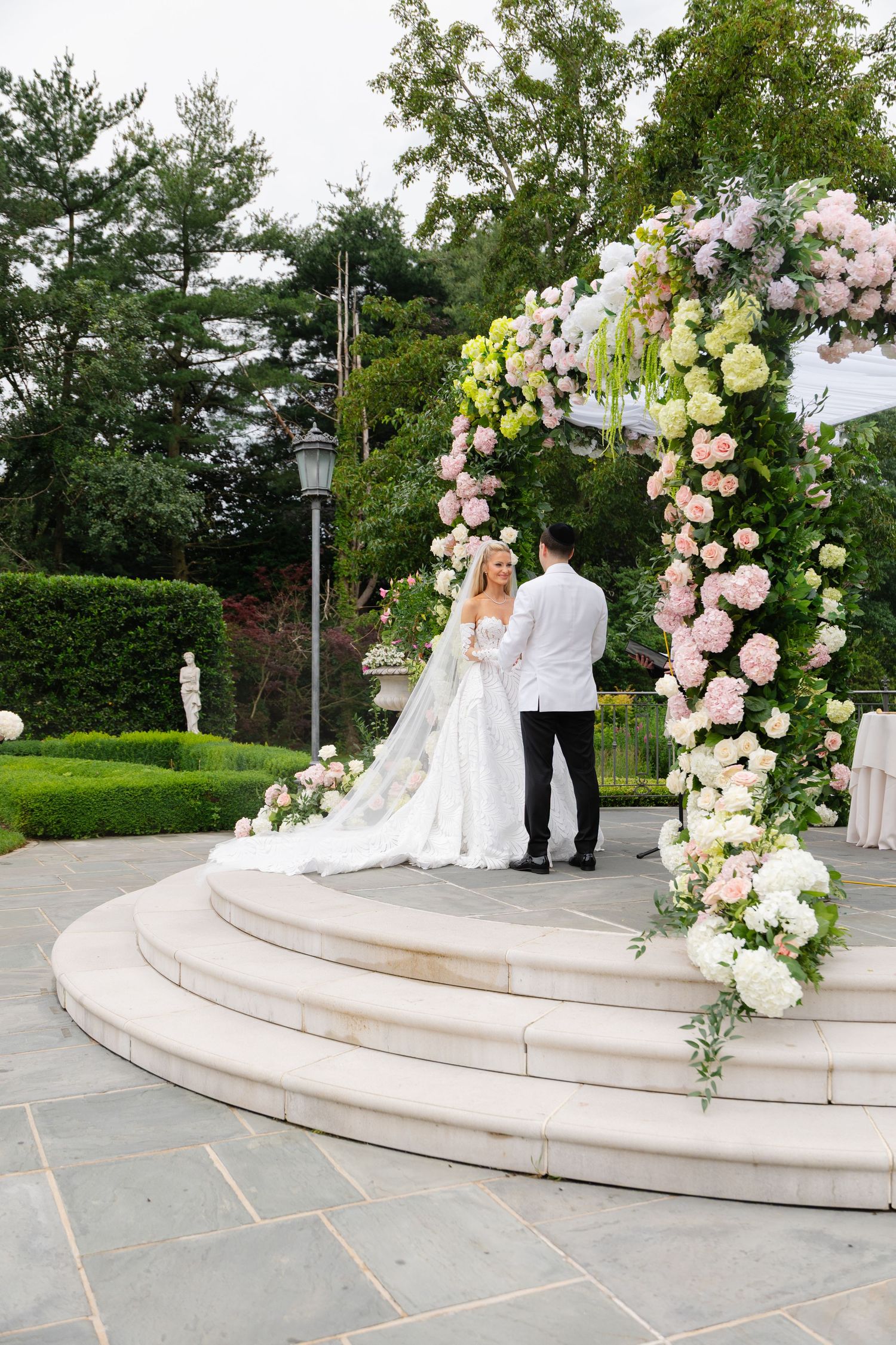 Bride and groom exchange vows under a floral arch adorned with pink and white roses in an elegant garden ceremony.