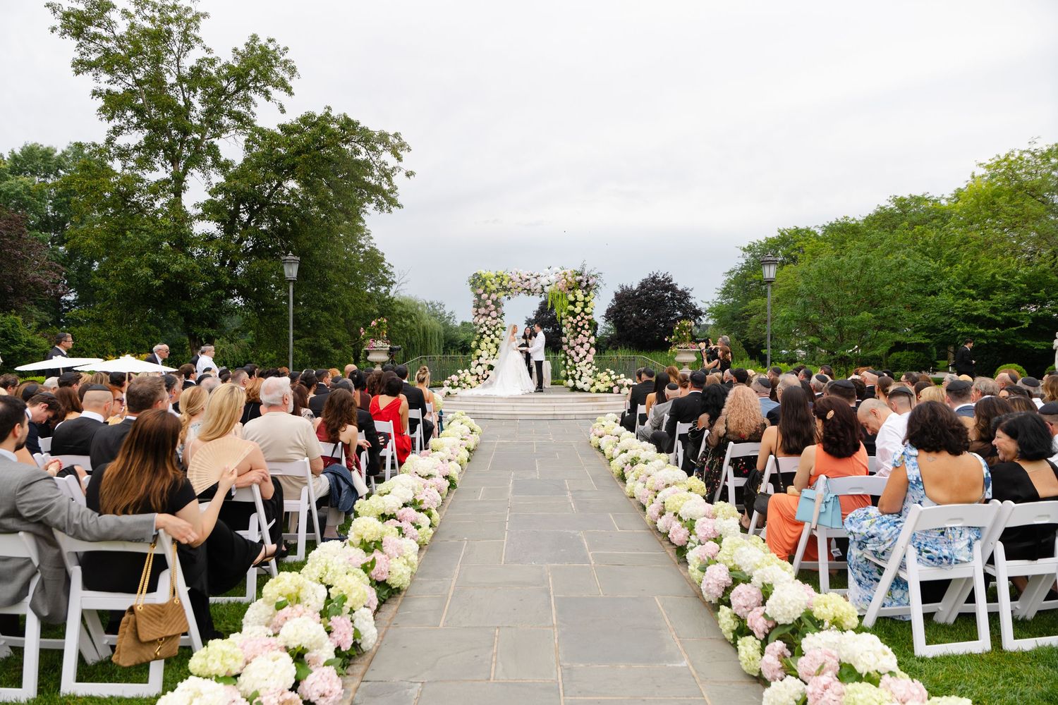 Outdoor wedding ceremony with floral aisle lined with pink hydrangeas, guests seated in white chairs, couple at flower arch.
