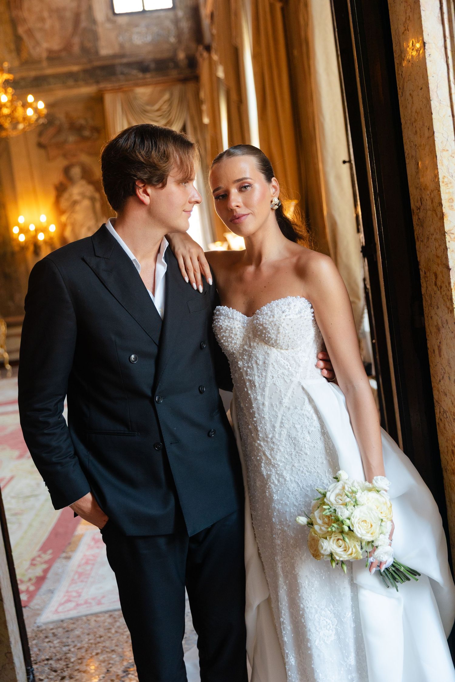 Elegant wedding couple in formal attire share intimate moments in an ornate hallway with chandeliers and gilded decor.