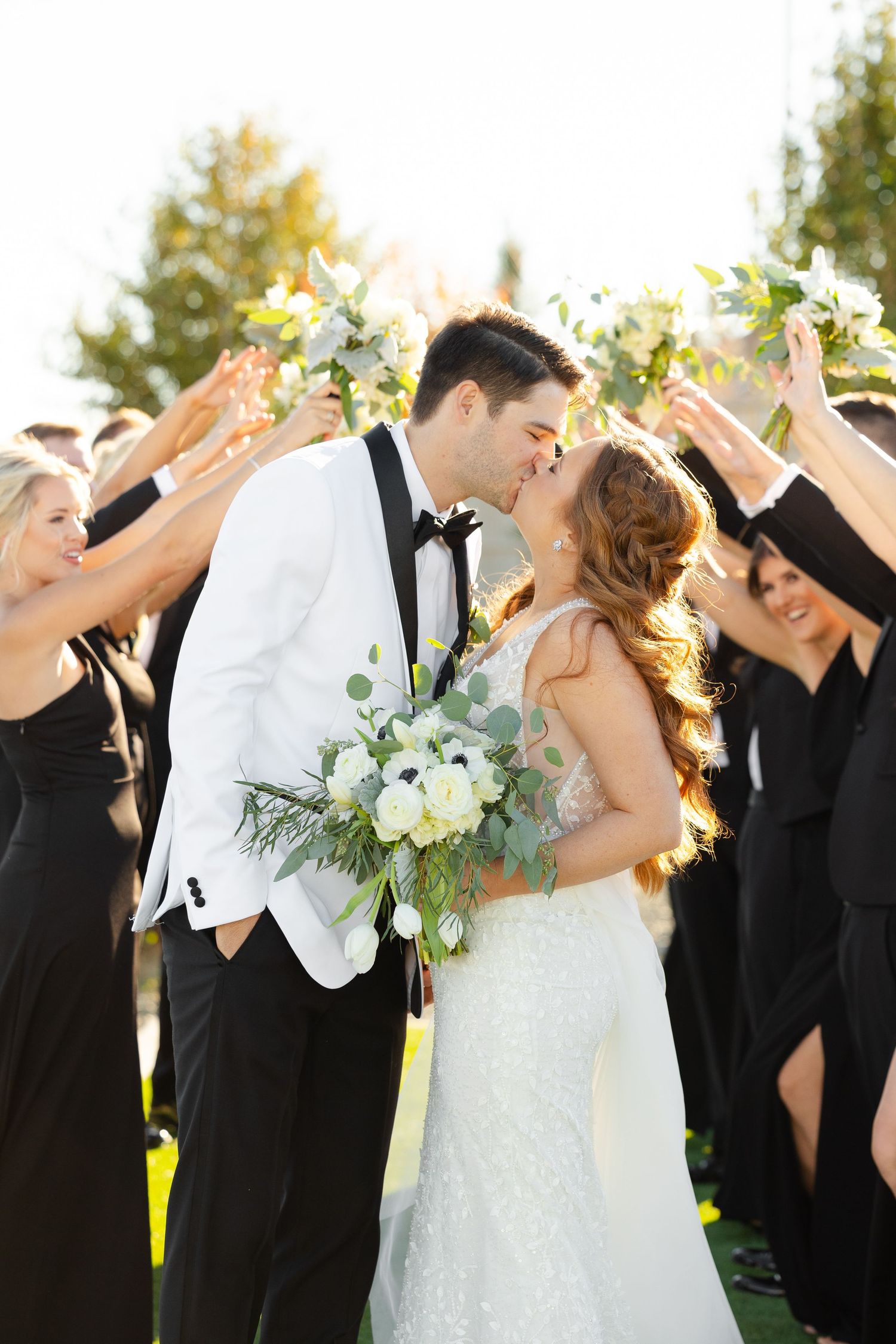 Wedding party in black and white attire poses outdoors with joyful expressions and floral bouquets.