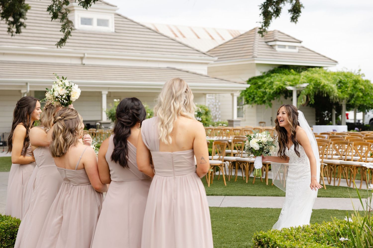 Bridesmaids in blush pink dresses walk together at an elegant outdoor wedding ceremony setup with wooden chairs.