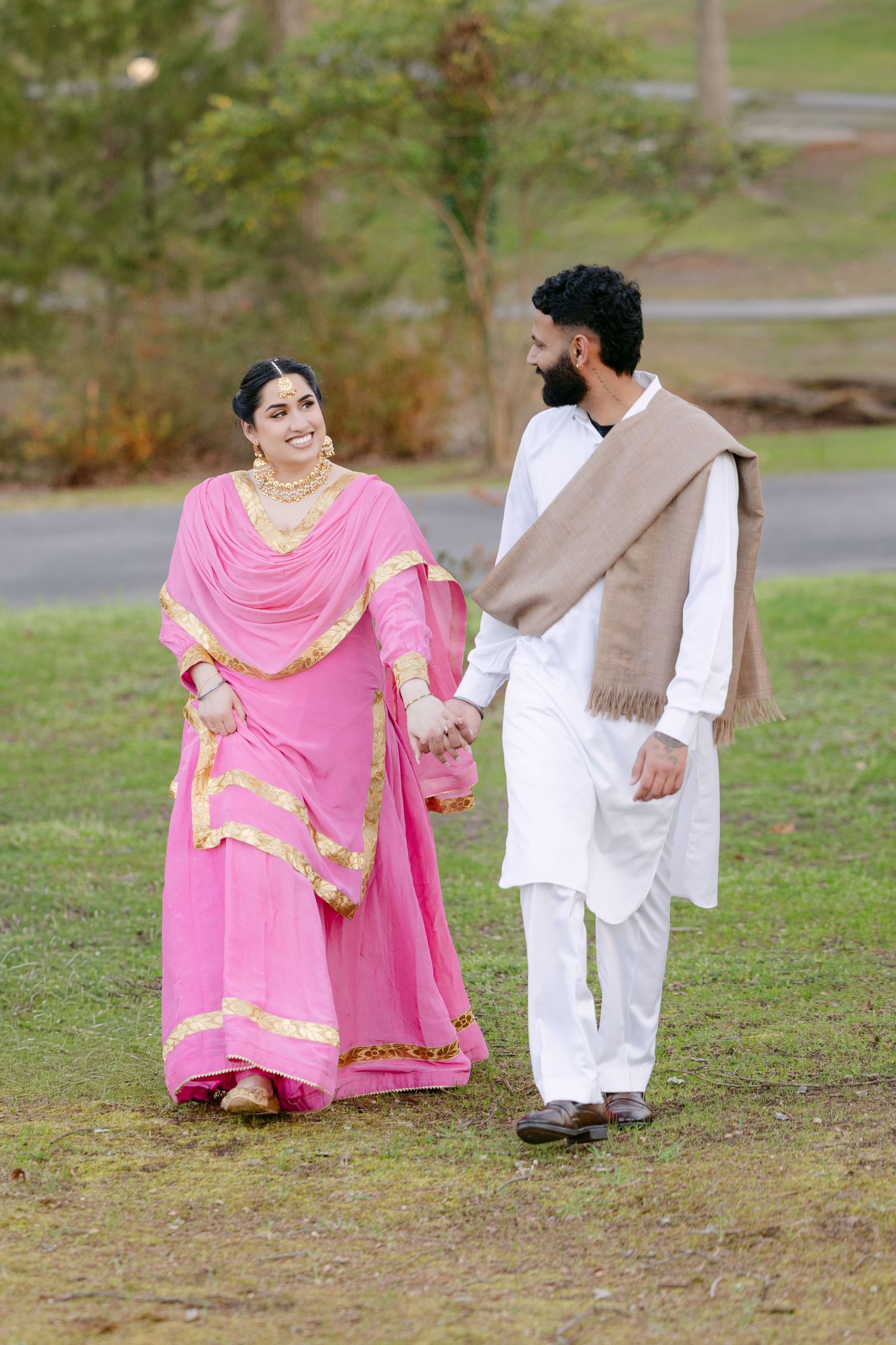 A couple in traditional South Asian attire, with pink and white clothing, walk together on grass in a park setting.