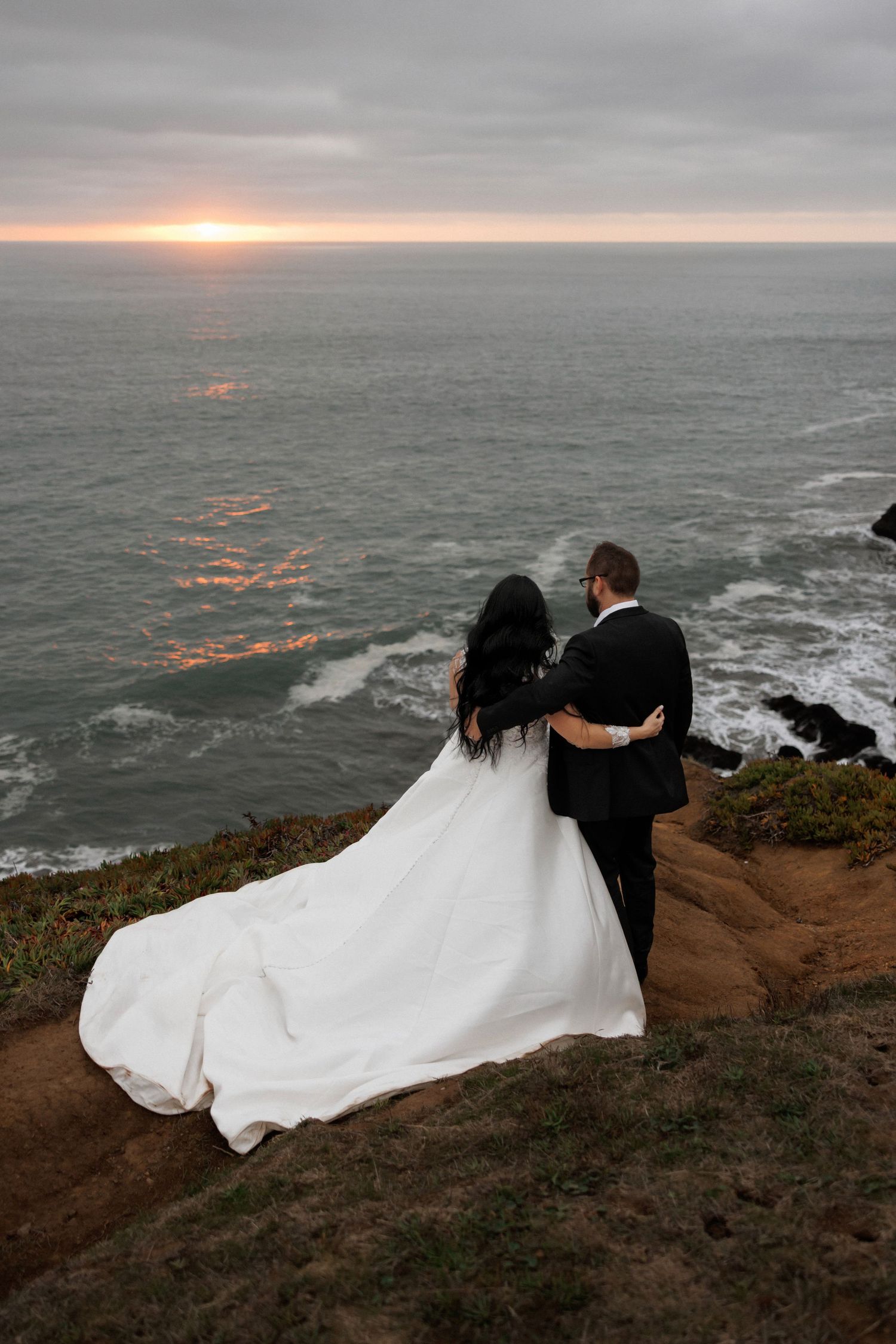 Dramatic cliffside wedding portraits overlooking ocean waves at sunset.