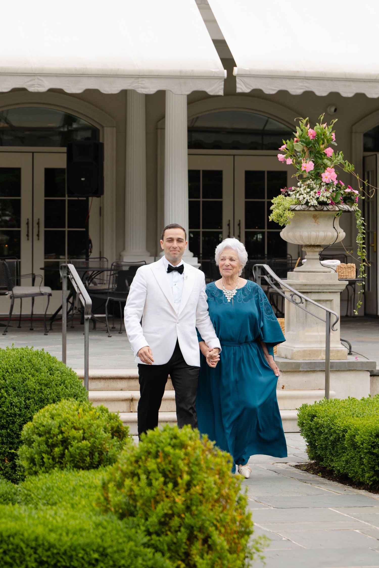 A groom in a white tuxedo and older woman in teal dress hold hands outside an elegant venue.
