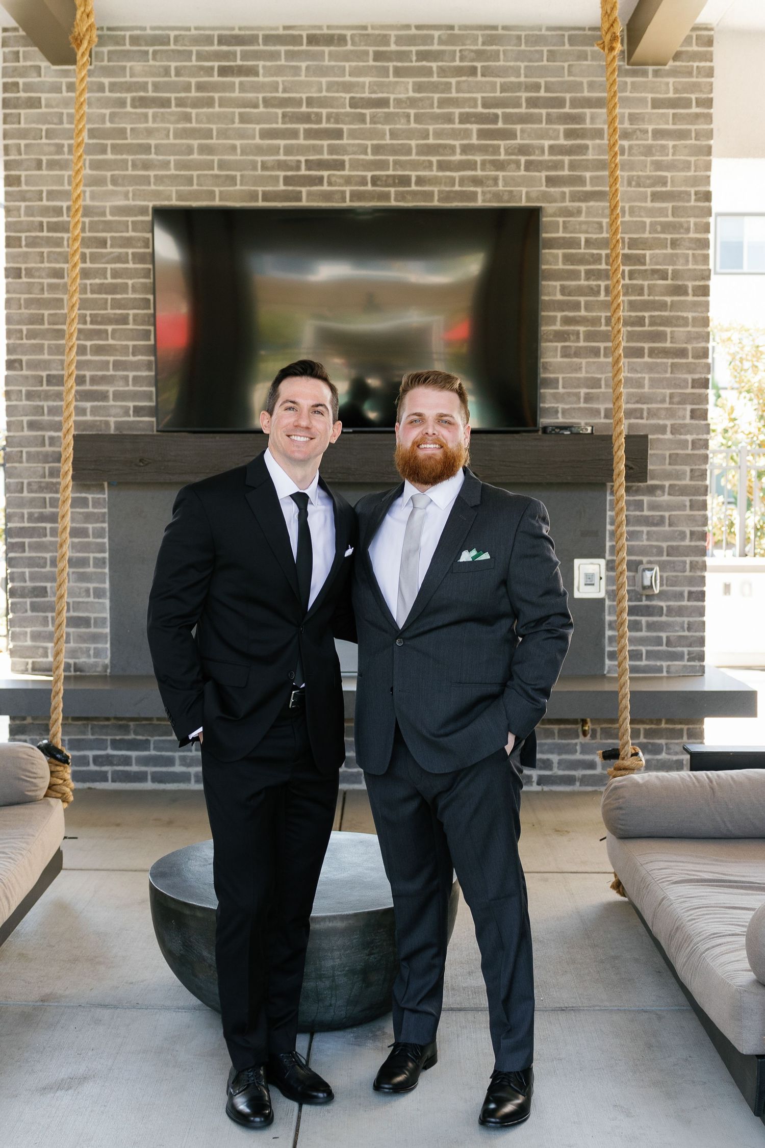 Two men in black suits and ties stand together in a modern lobby with brick walls and leather furniture.