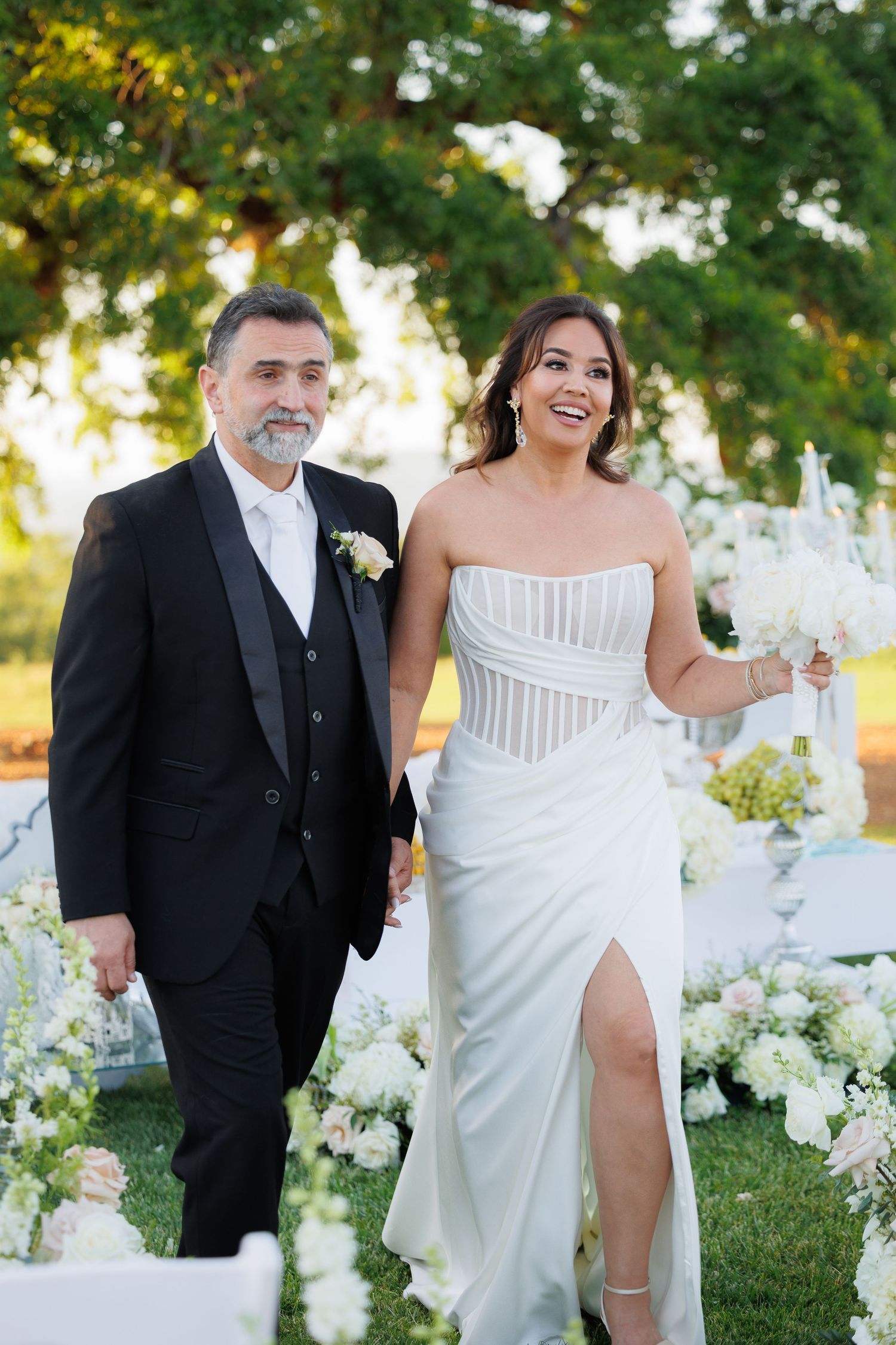Couple in formal wedding attire walk and laugh together in a lush garden setting with white floral decorations.
