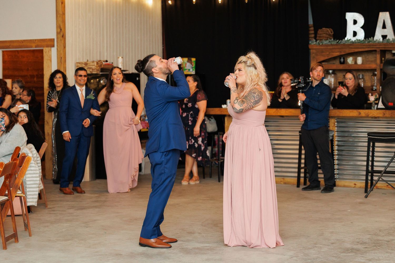 Wedding party members in pink and blue formal attire dance together at a rustic indoor reception venue.