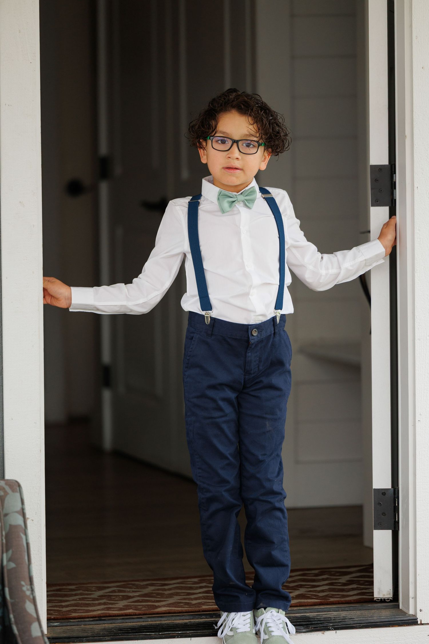 A young child in a white shirt, navy suspenders and pants leans against a white porch column in a casual pose.
