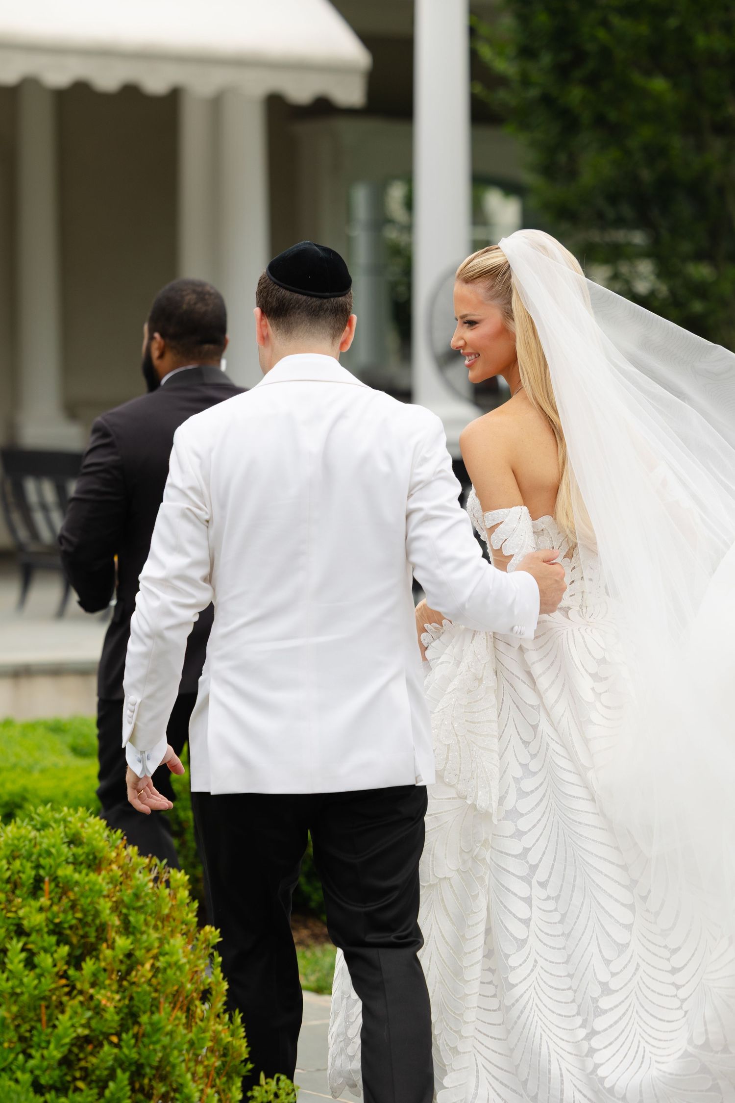 A bride and groom share a candid moment together in front of white columns and green landscaping.