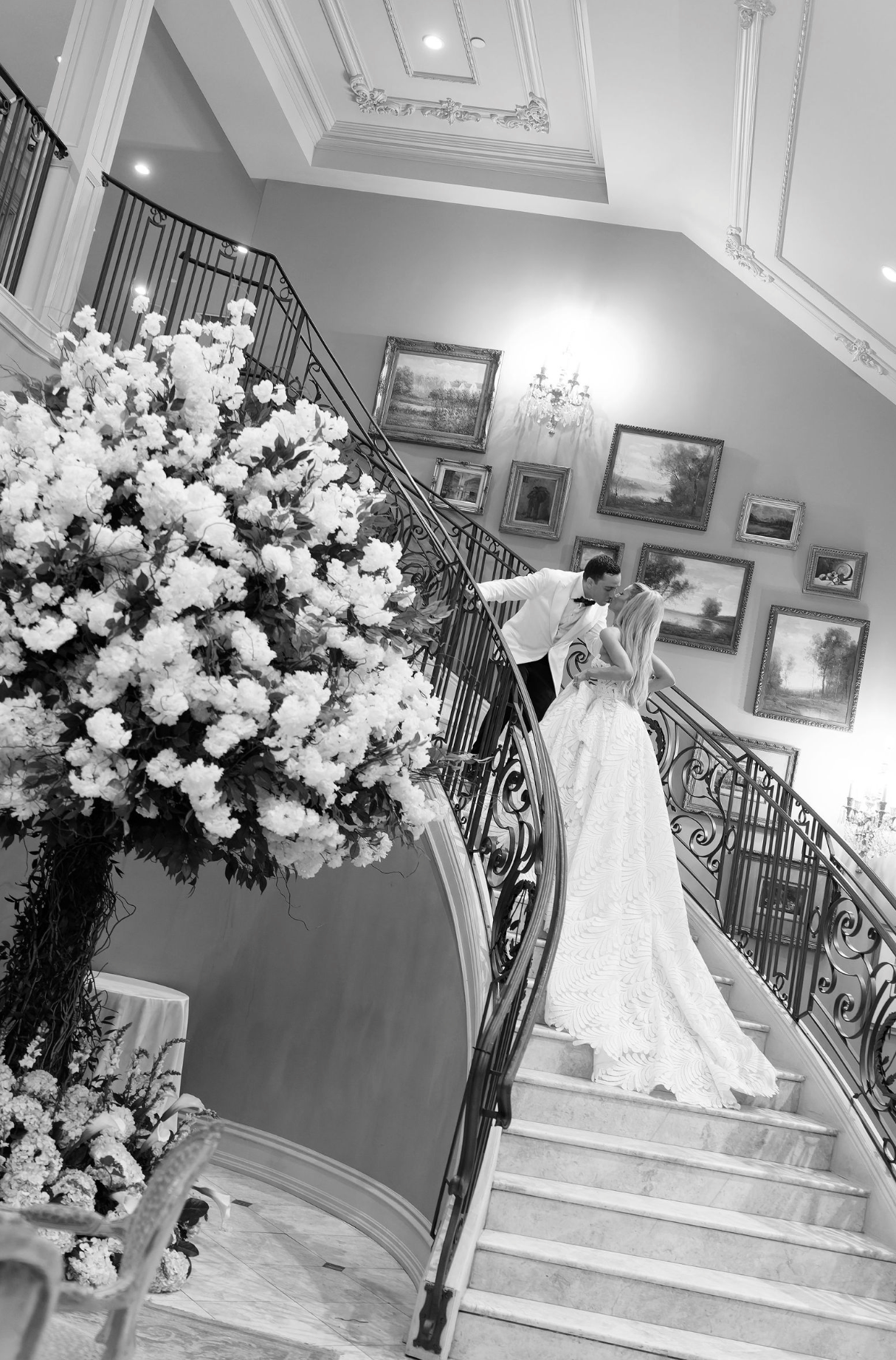 A wedding dress displayed on a grand staircase with ornate railings and floral arrangements in an elegant venue.