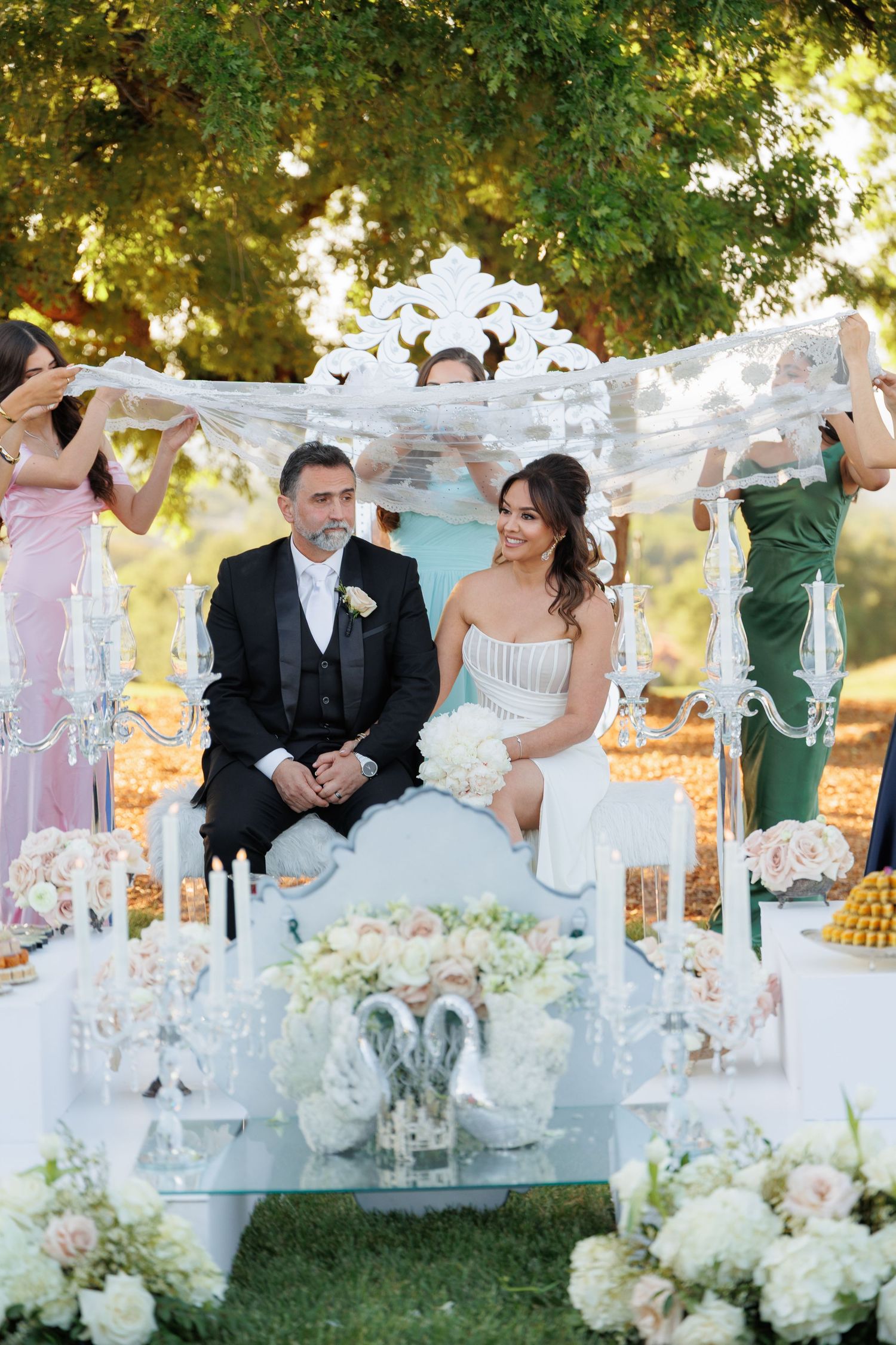 Elegant outdoor wedding reception setup with white floral centerpieces and delicate lace decorations hanging above.