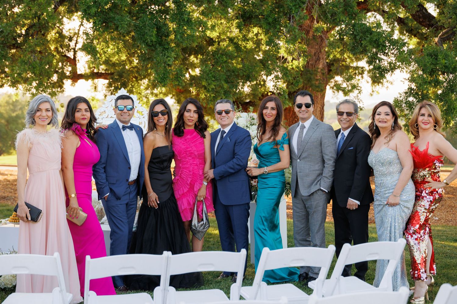 A group in formal attire poses together at an outdoor wedding venue with white chairs and trees in the background.