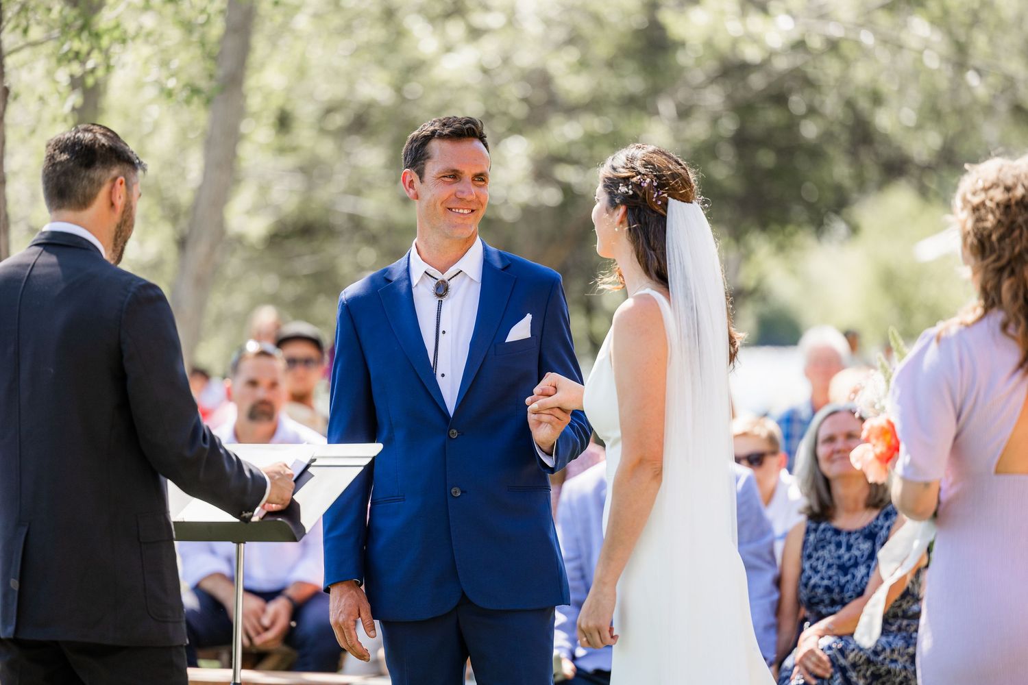 A wedding ceremony takes place outdoors with the couple exchanging vows in front of guests on a sunny day.