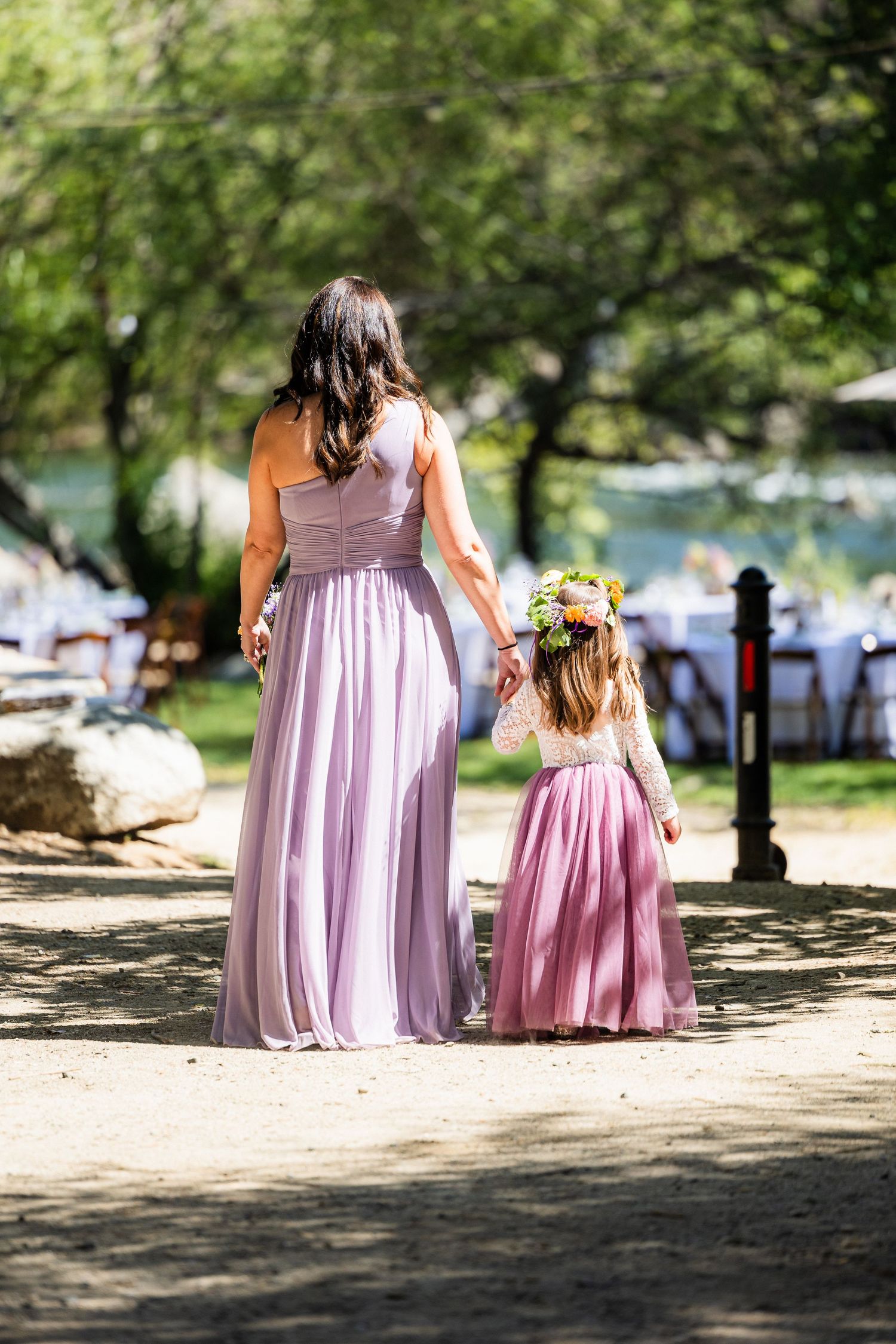 Two figures in matching pink dresses walk down a path at an outdoor wedding venue beside a lake.