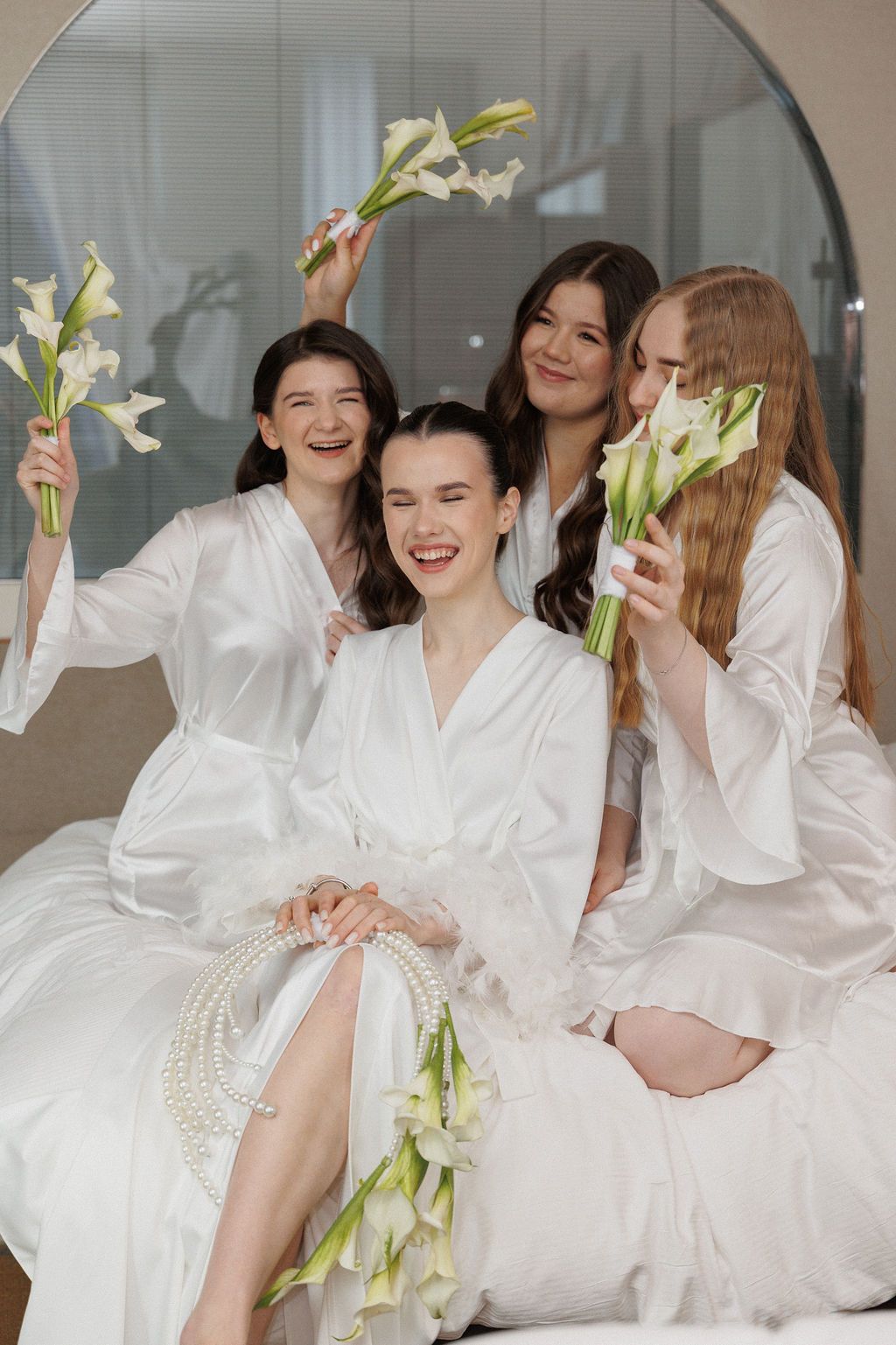 A group wearing white robes and holding calla lilies laughs together during a pre-wedding celebration.