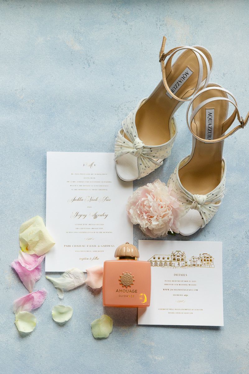 Wedding details flatlay with elegant shoes, invitations, and rose petals on a light blue background.