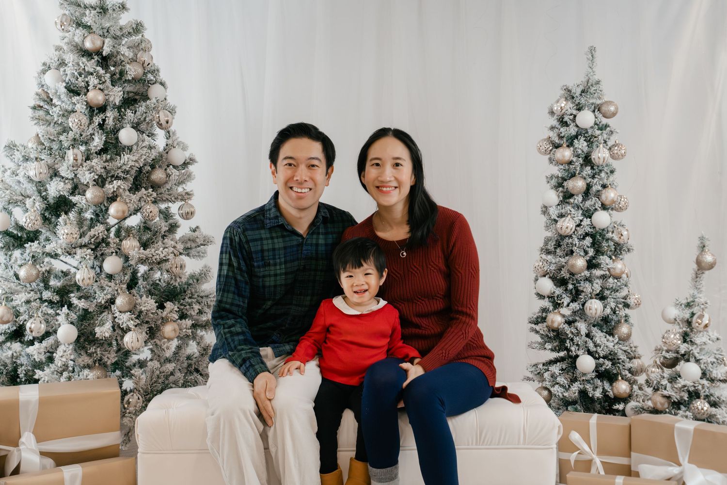 Mom, Dad, Kid smiling together on a white bench between snowy Christmas trees during an indoor Christmas family photoshoot in Kingston, On