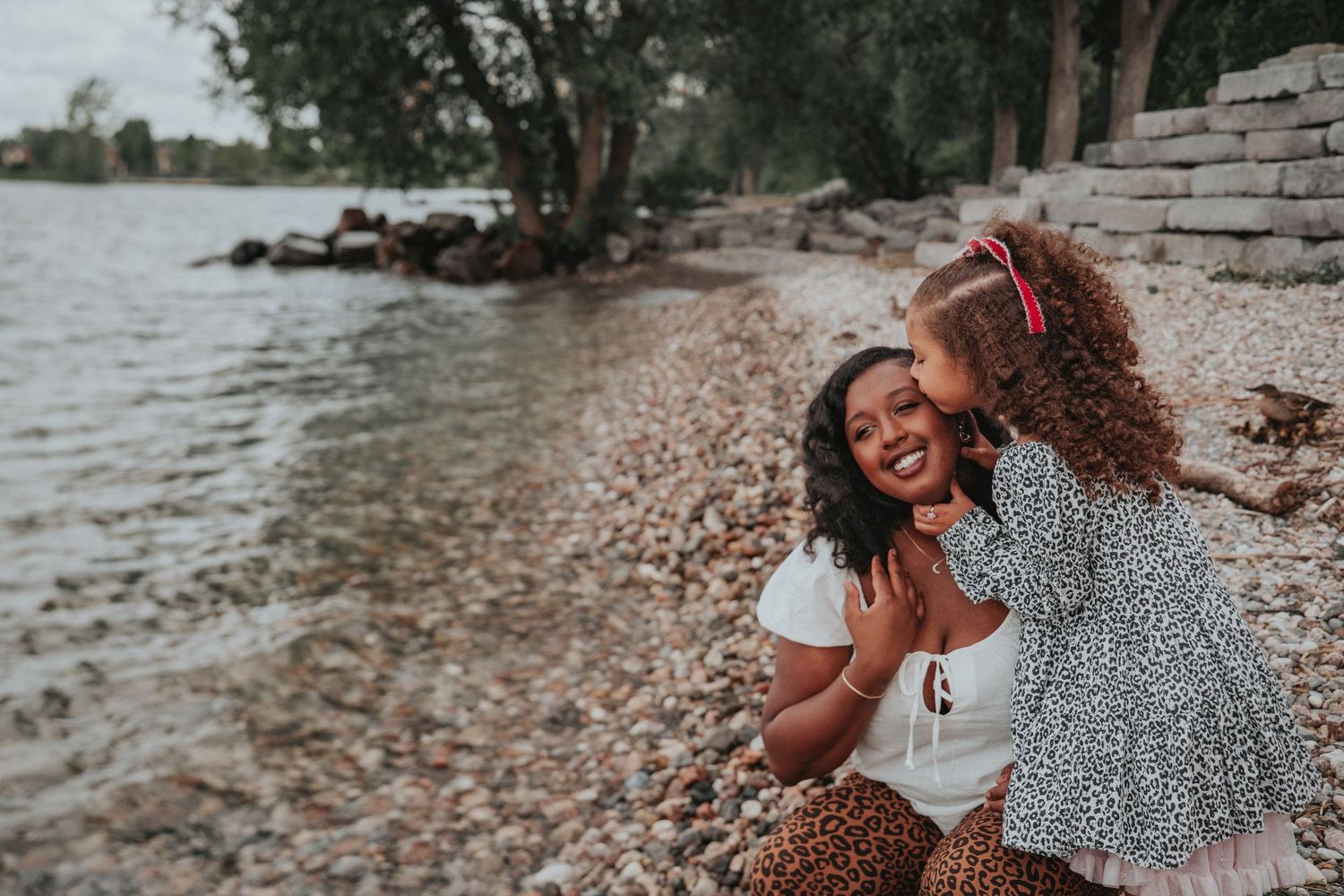 Mother hugging her smiling daughter along a rocky waterfront during a candid outdoor summer family photography session in Kingston, Ontario.