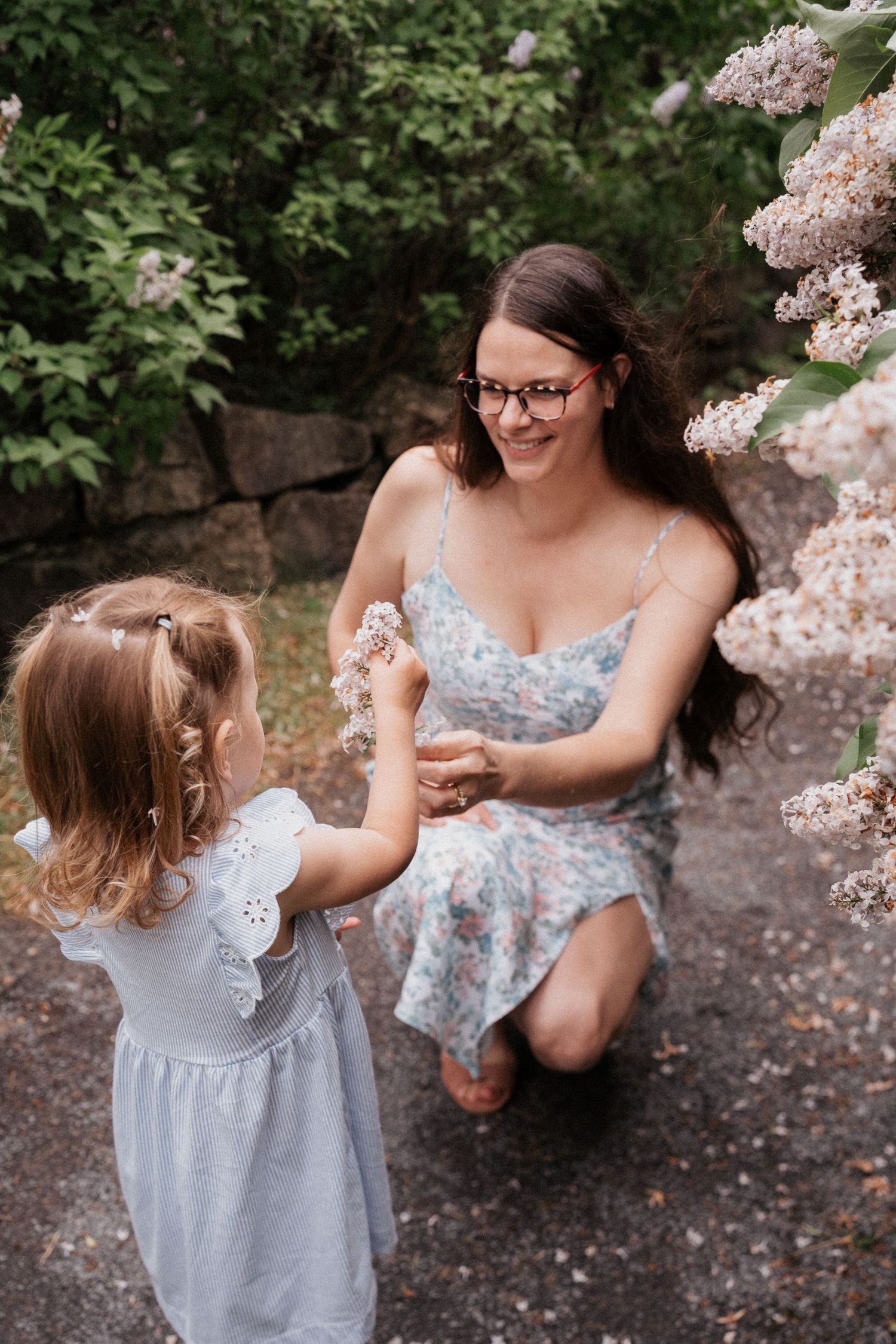 Mother kneeling beside her young daughter on a garden path during a spring family photography session surrounded by blooming flowers.
