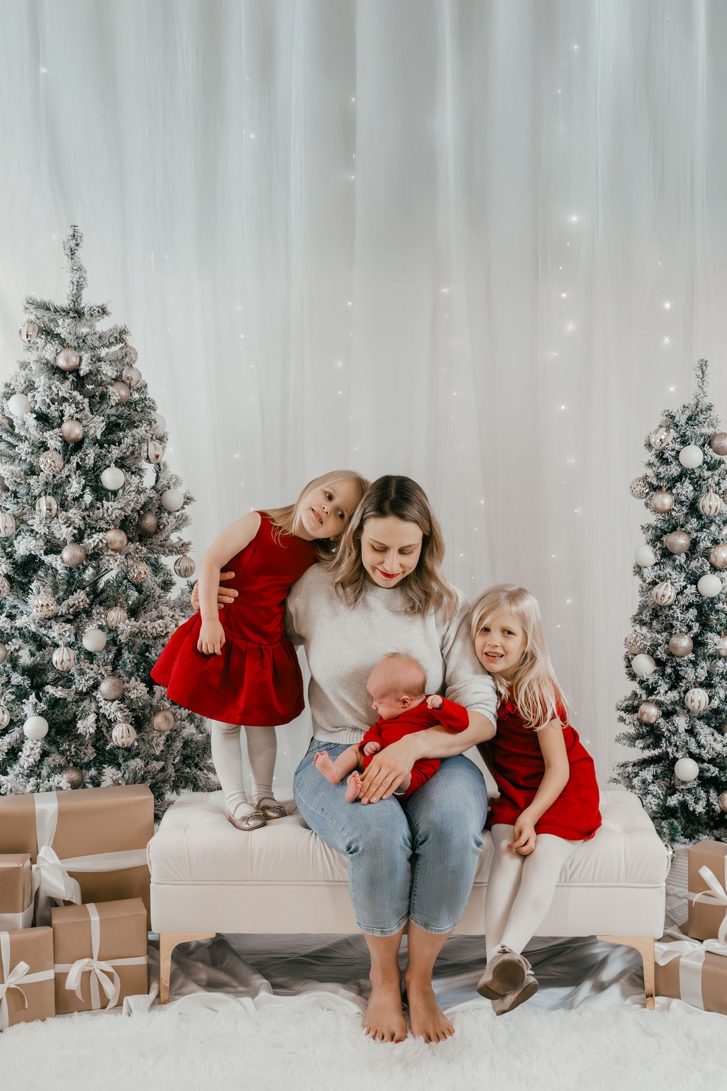 Christmas mini session featuring a mom and daughters seated together on a festive indoor holiday backdrop with Christmas trees in Kingston