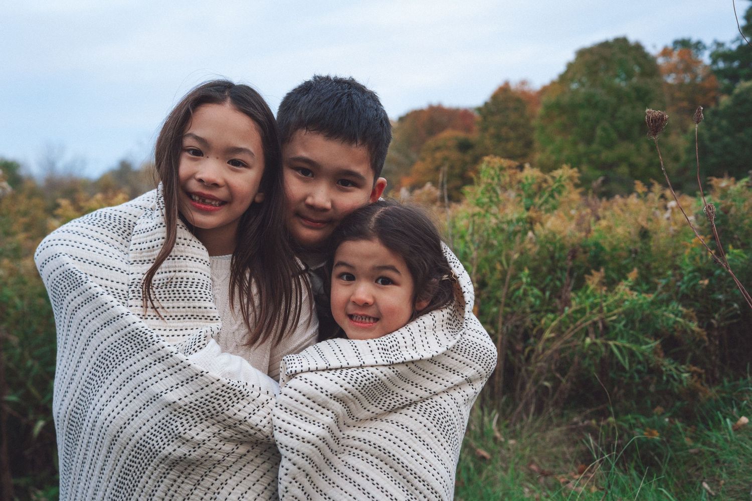 Family of sisters and brother smiling and cuddling together outdoors during a fall family photography session in Kingston, Ontario.