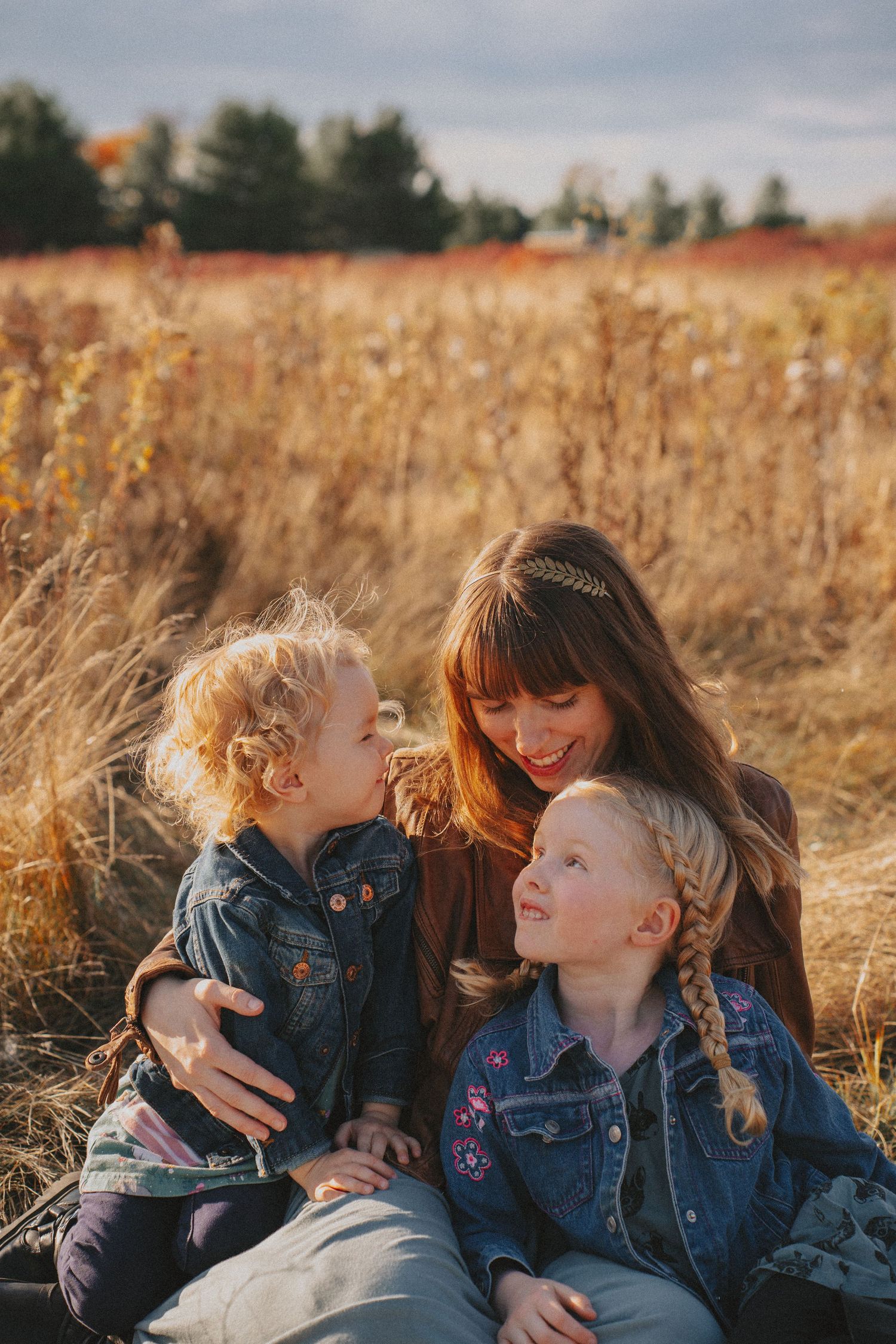 Mother cuddling her two young children in a golden grassy field during an outdoor fall family portrait session in Kingston, Ontario.