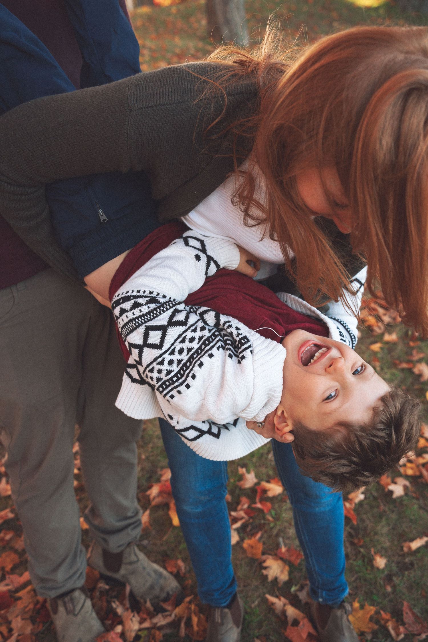 Mother leaning down to kiss her young child during a candid outdoor fall family photography session surrounded by autumn leaves.