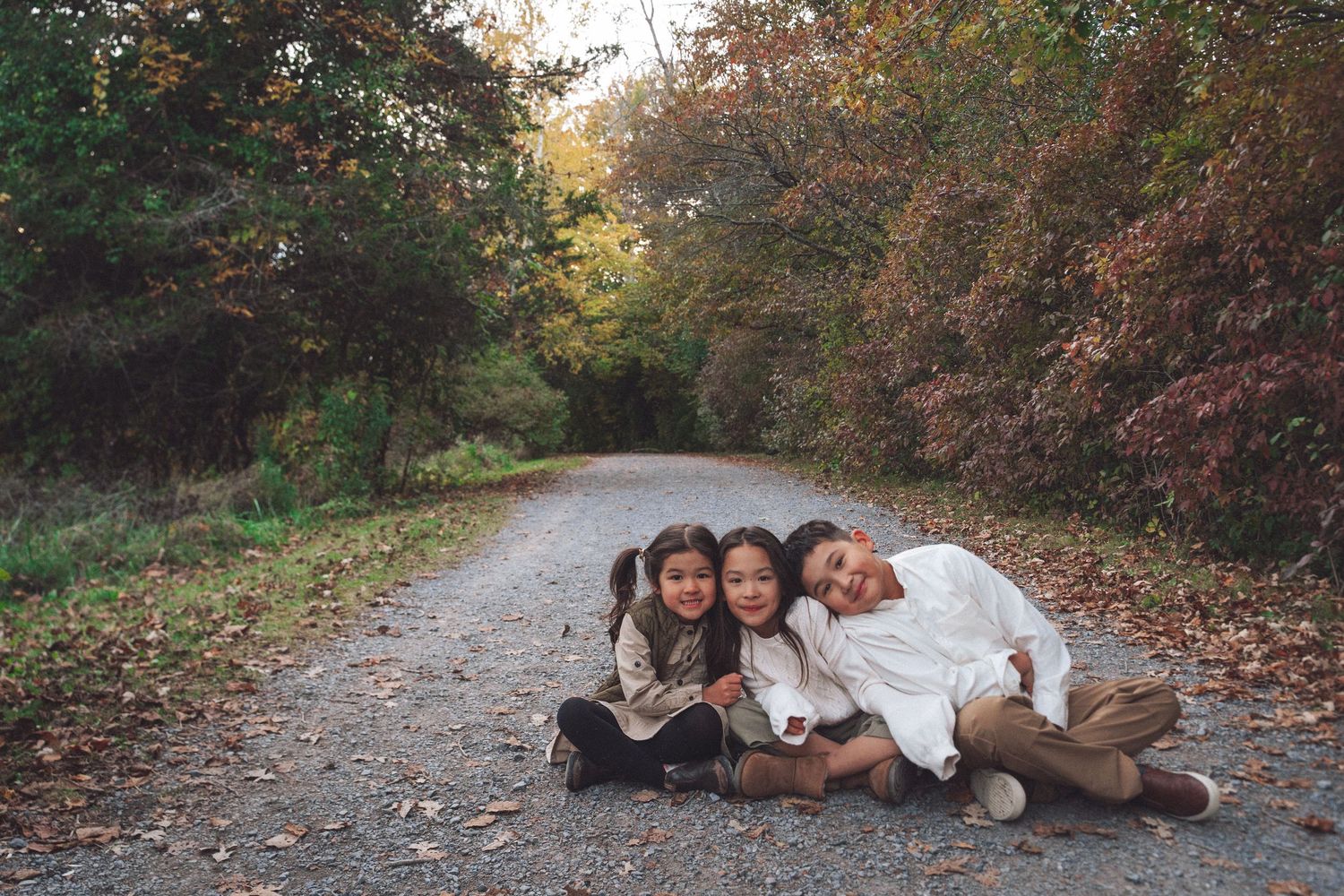 Family of three sitting together on a tree-lined path surrounded by autumn foliage during an outdoor family photoshoot in Kingston, Ontario.
