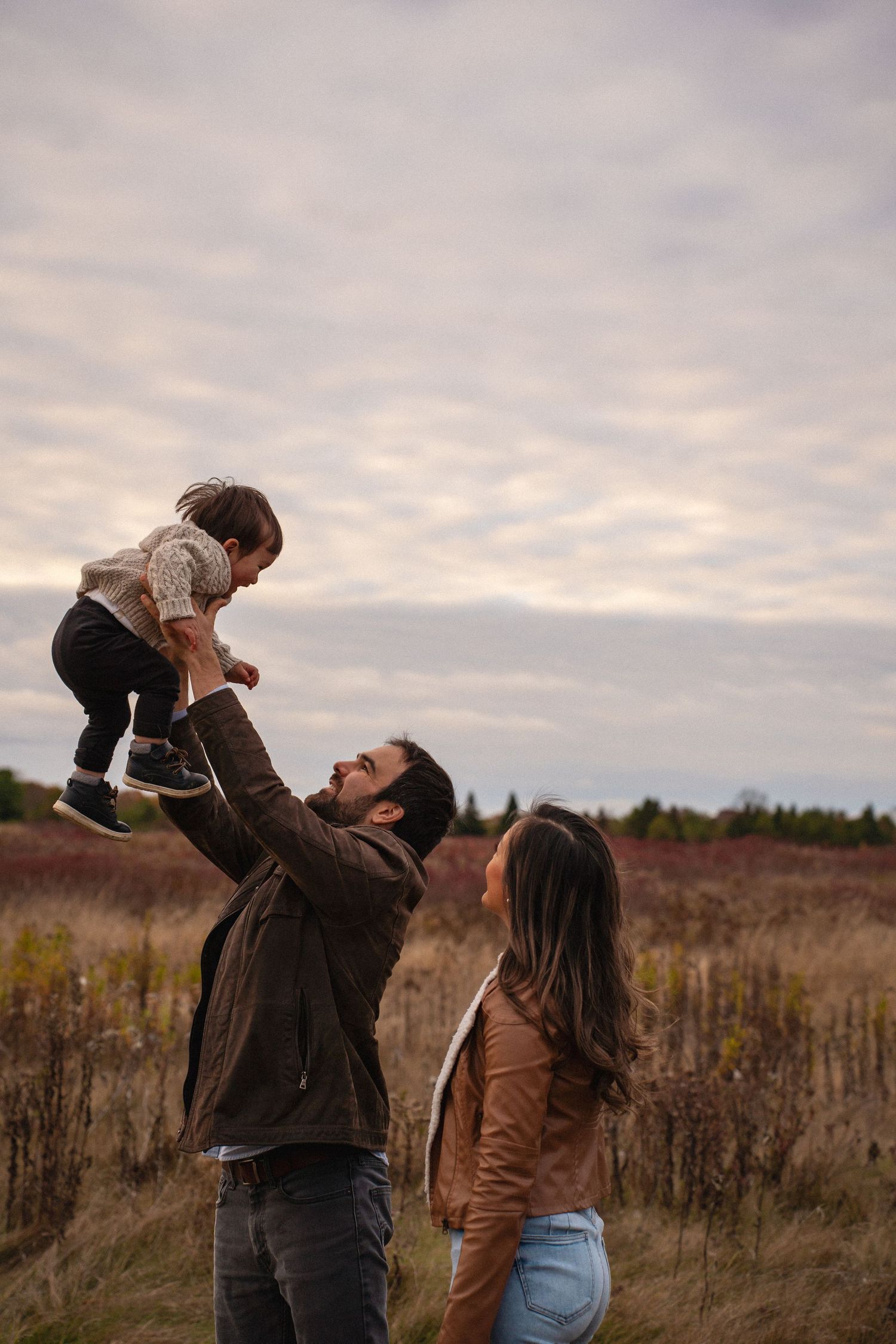 Father lifting his young child into the air while the mom watches during an outdoor fall family photoshoot in Lemoine Pt Kingston, Ontario.