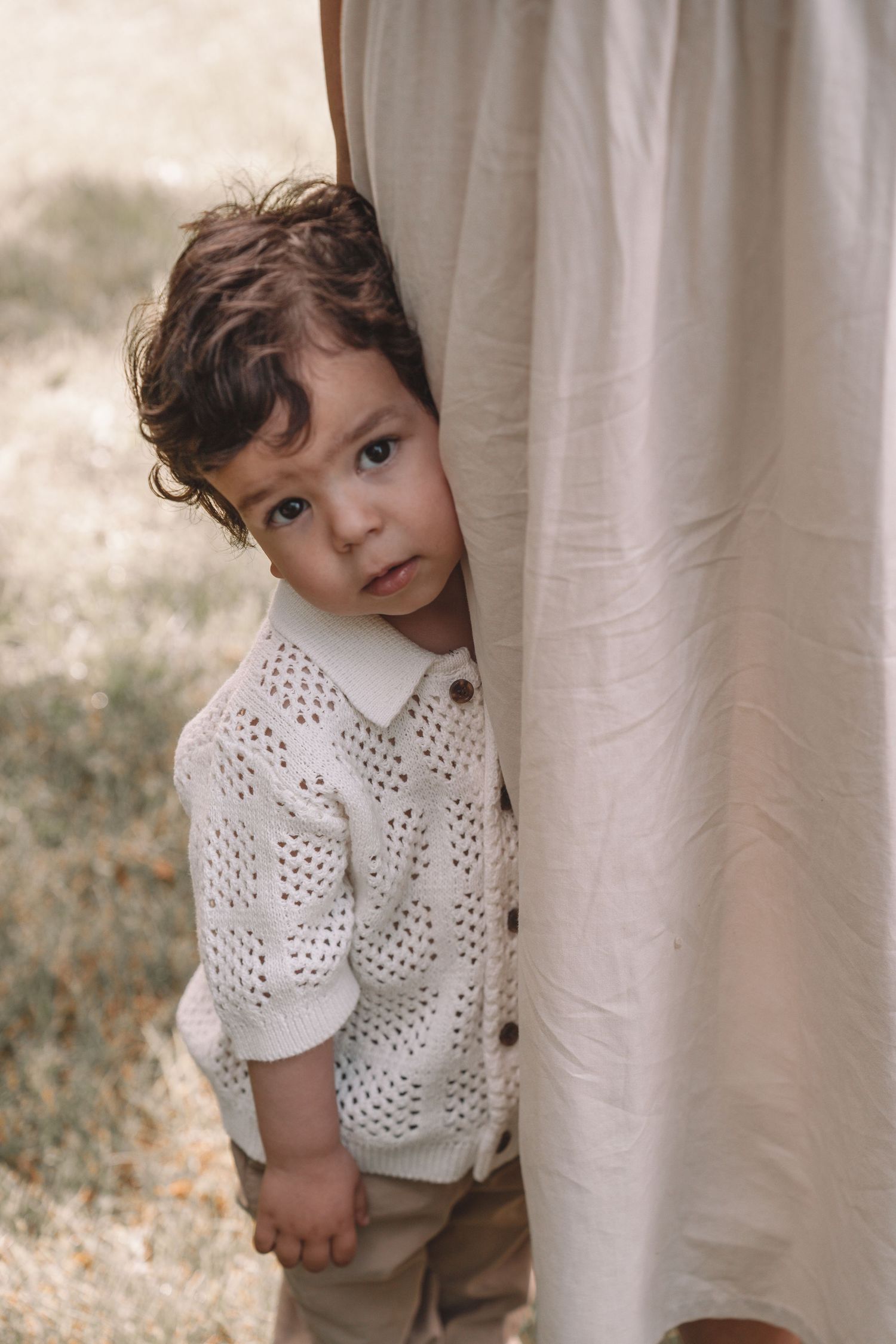 Young boy toddler peeking out from behind his parent's leg during an outdoor family photography session in Kingston, Ontario.