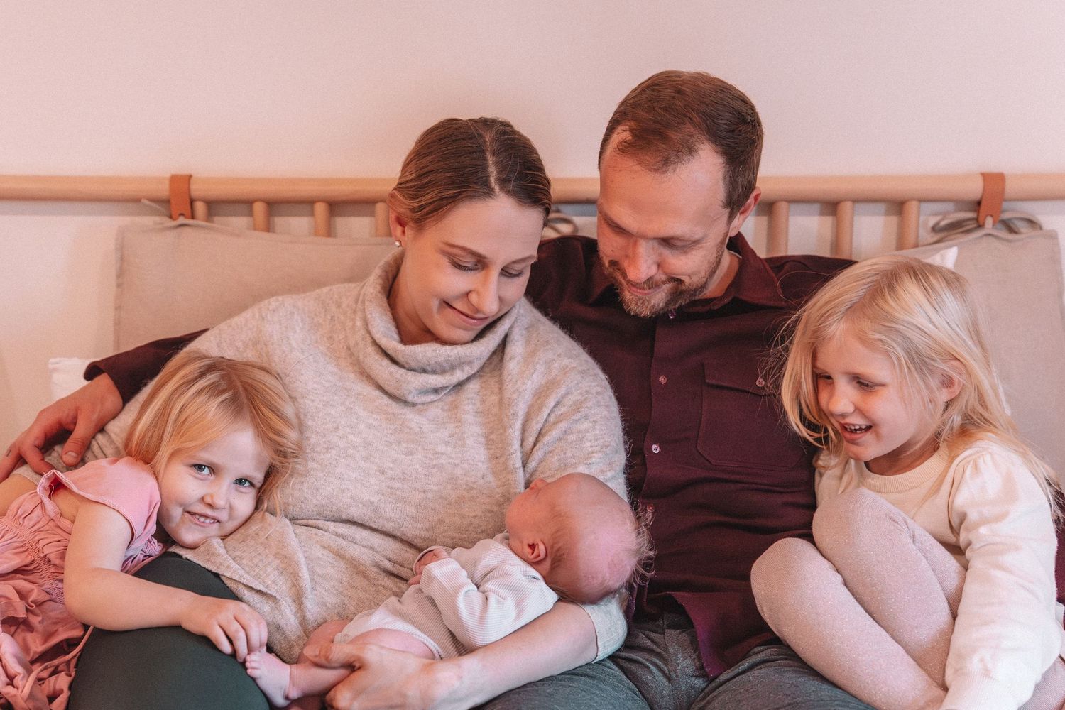 Parents cuddling their newborn baby on a bed with two young daughters during a cozy, at-home newborn family photography session.