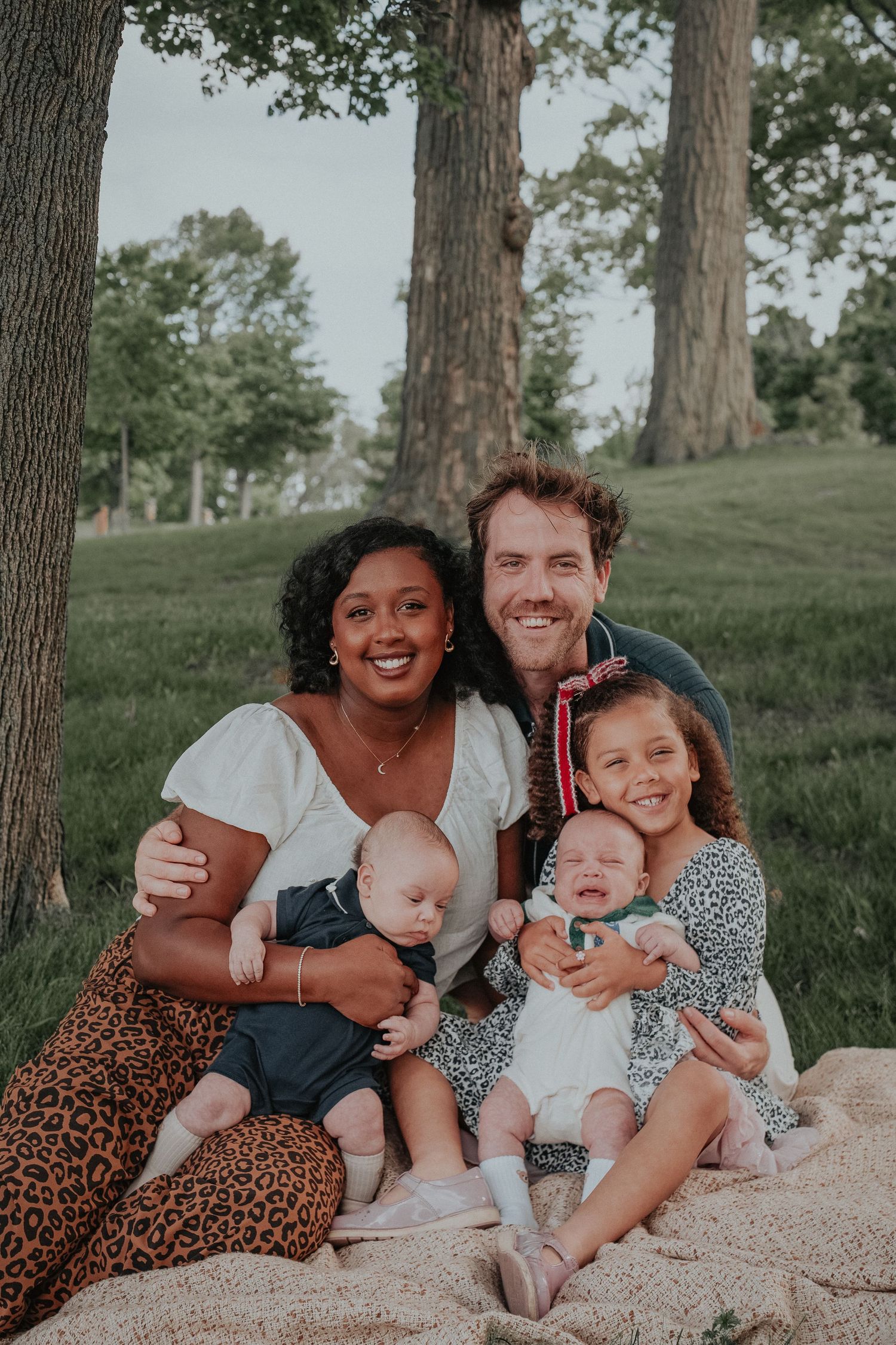 Family of four sitting together beneath tall trees during a relaxed outdoor summer family portrait session in Kingston, Ontario.