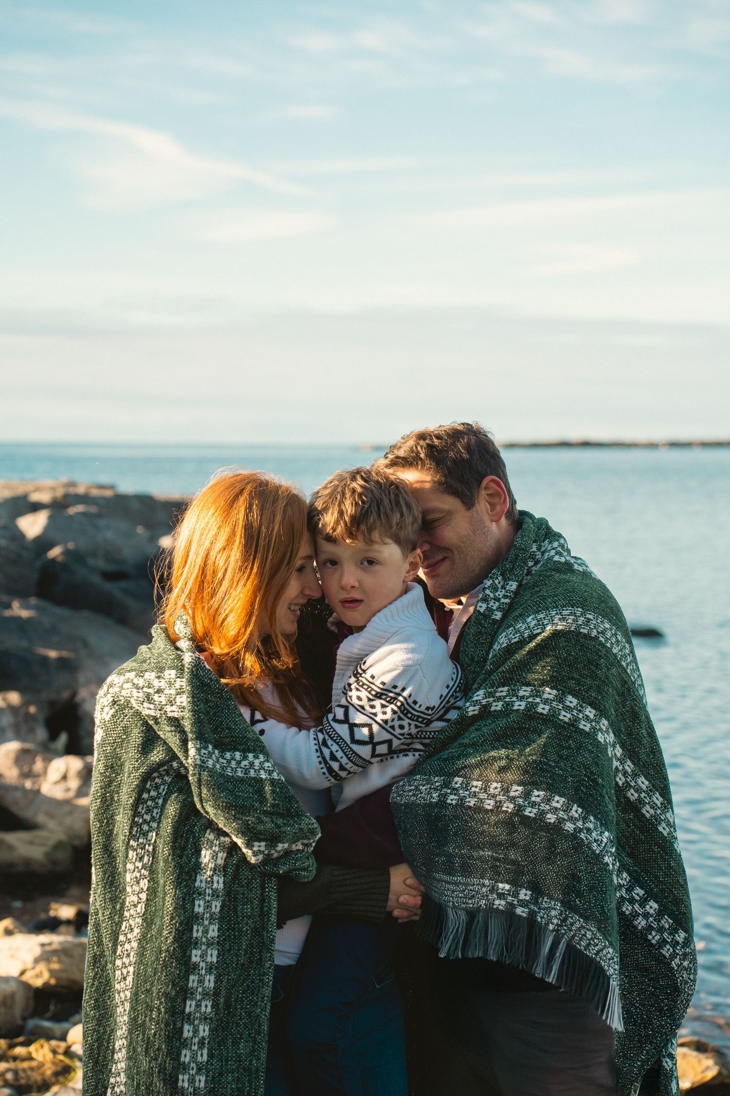 Parents embracing their young child by the Lake Ontario Park waterfront during a candid outdoor family photo session in Kingston, Ontario.