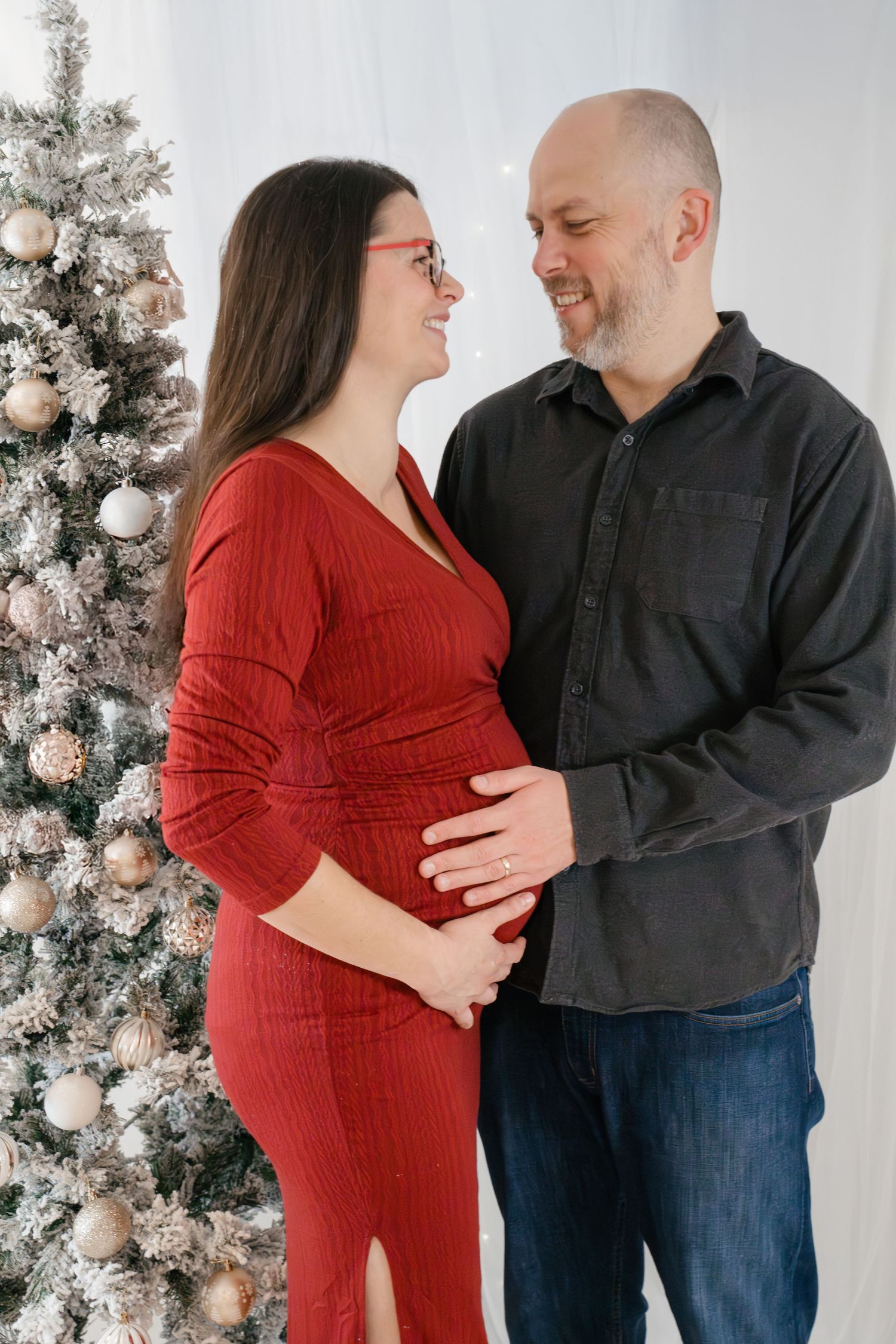 Expecting pregnant couple posing for a Christmas maternity photoshoot beside a snowy Christmas tree at Twy Photography in Kingston, Ontario.