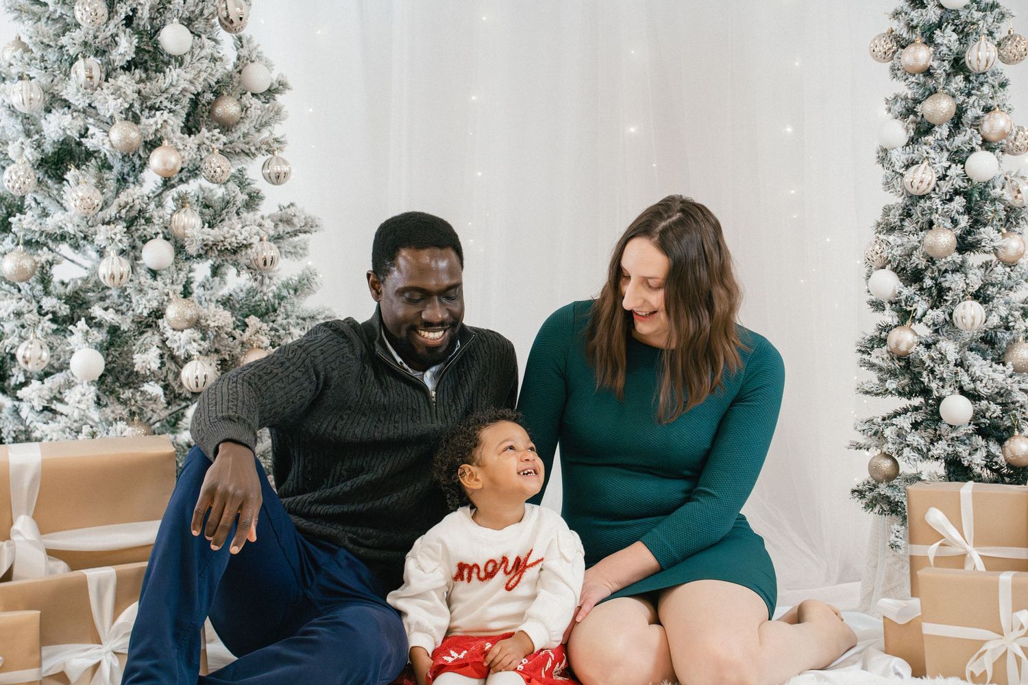 Family of three smiling together on a festive indoor Christmas backdrop with snowy trees during a holiday mini session in Kingston, Ontario.