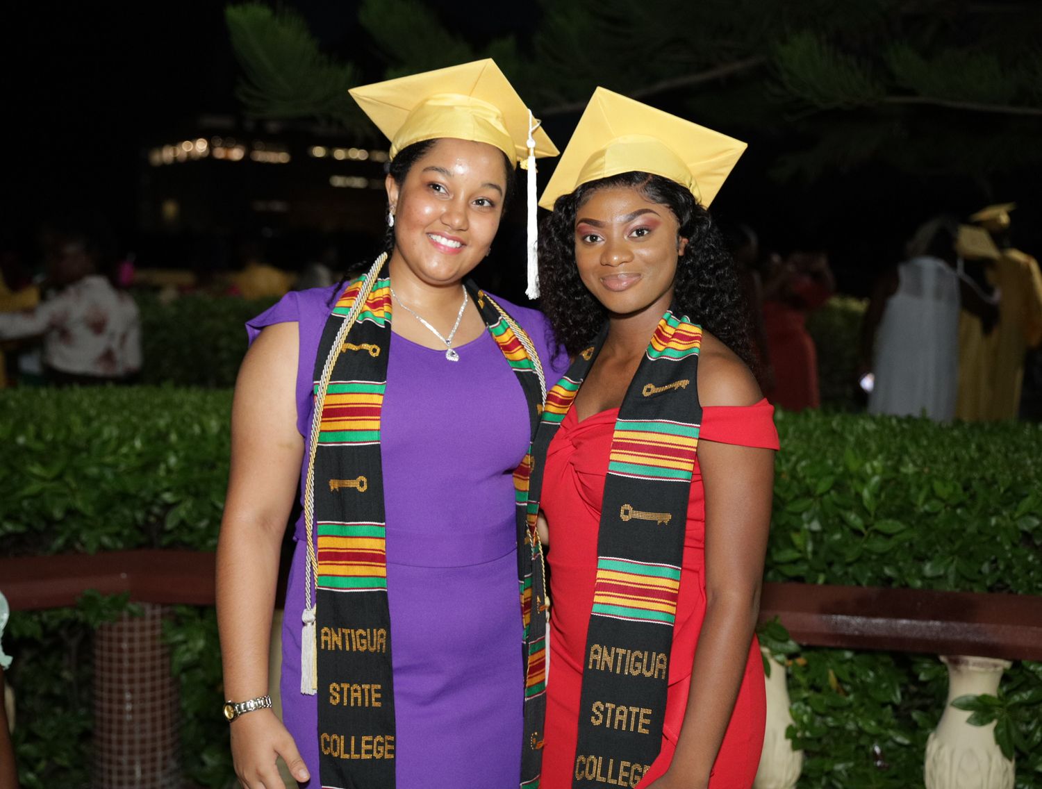 Two smiling graduates in gold caps and colorful Antigua State College stoles pose together at a graduation ceremony.