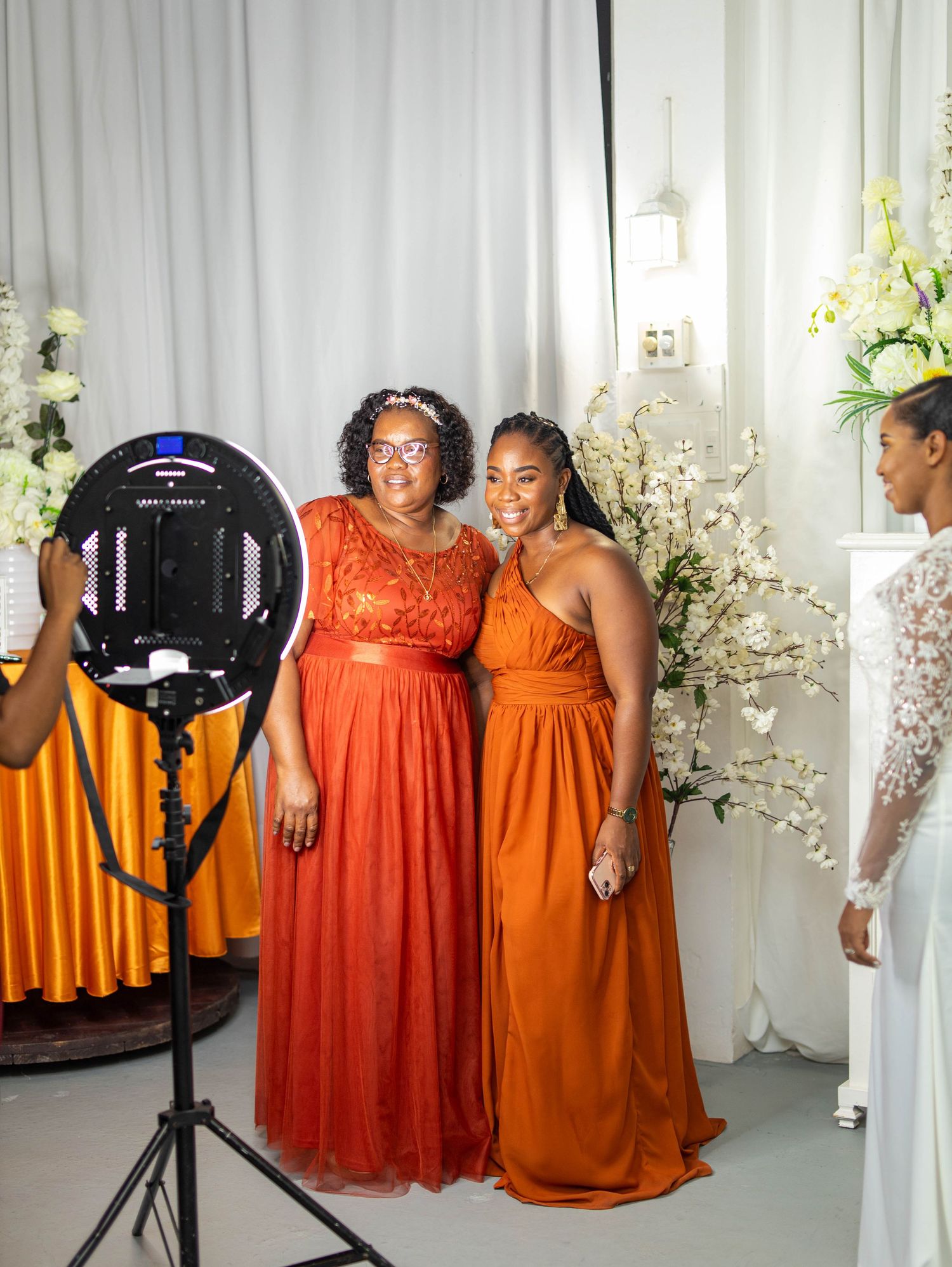 A group of bridesmaids in vibrant orange and coral dresses pose together in a wedding venue with white floral decor.