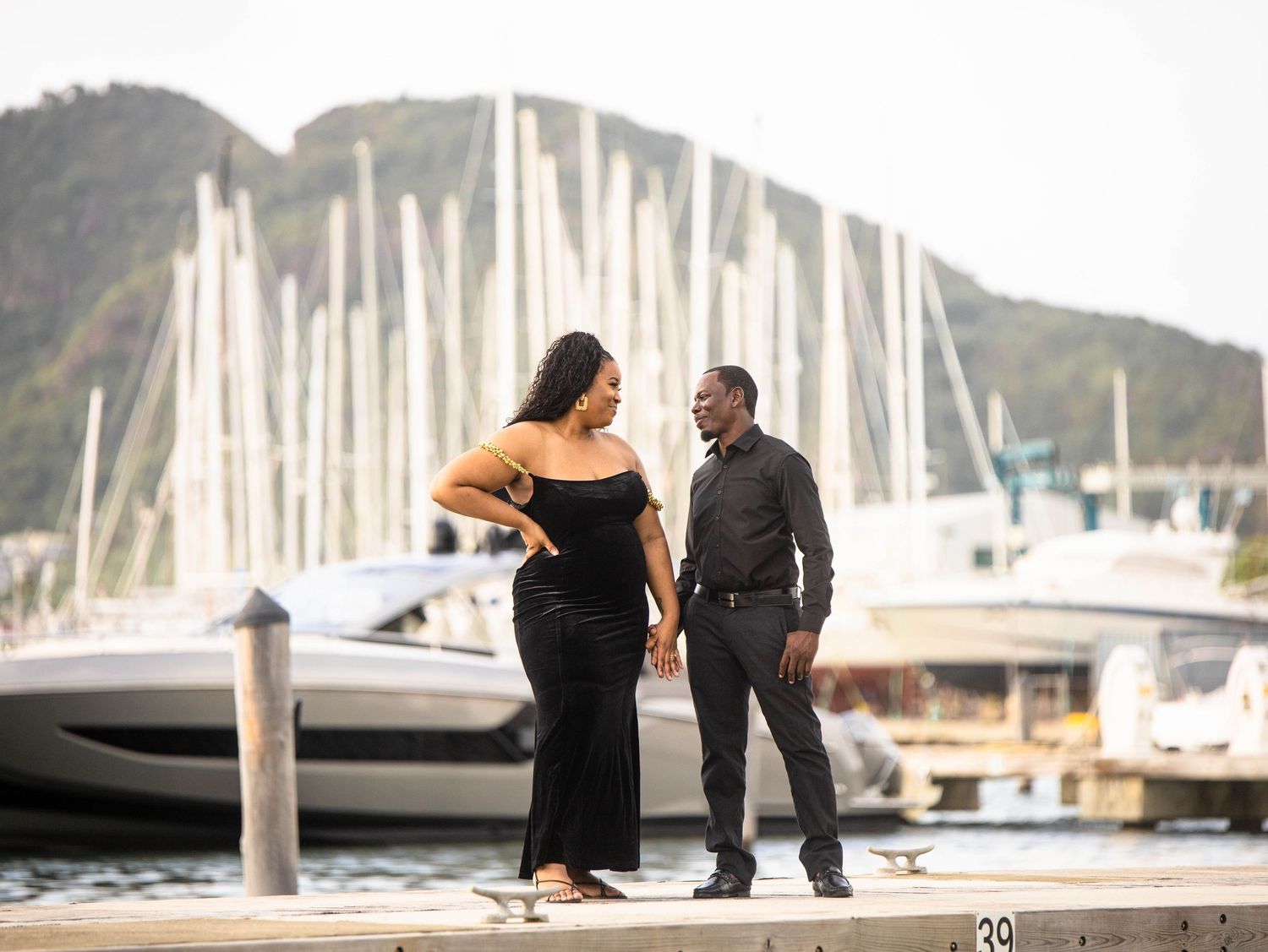 A couple stands on a marina dock with sailboat masts and mountains in the background.