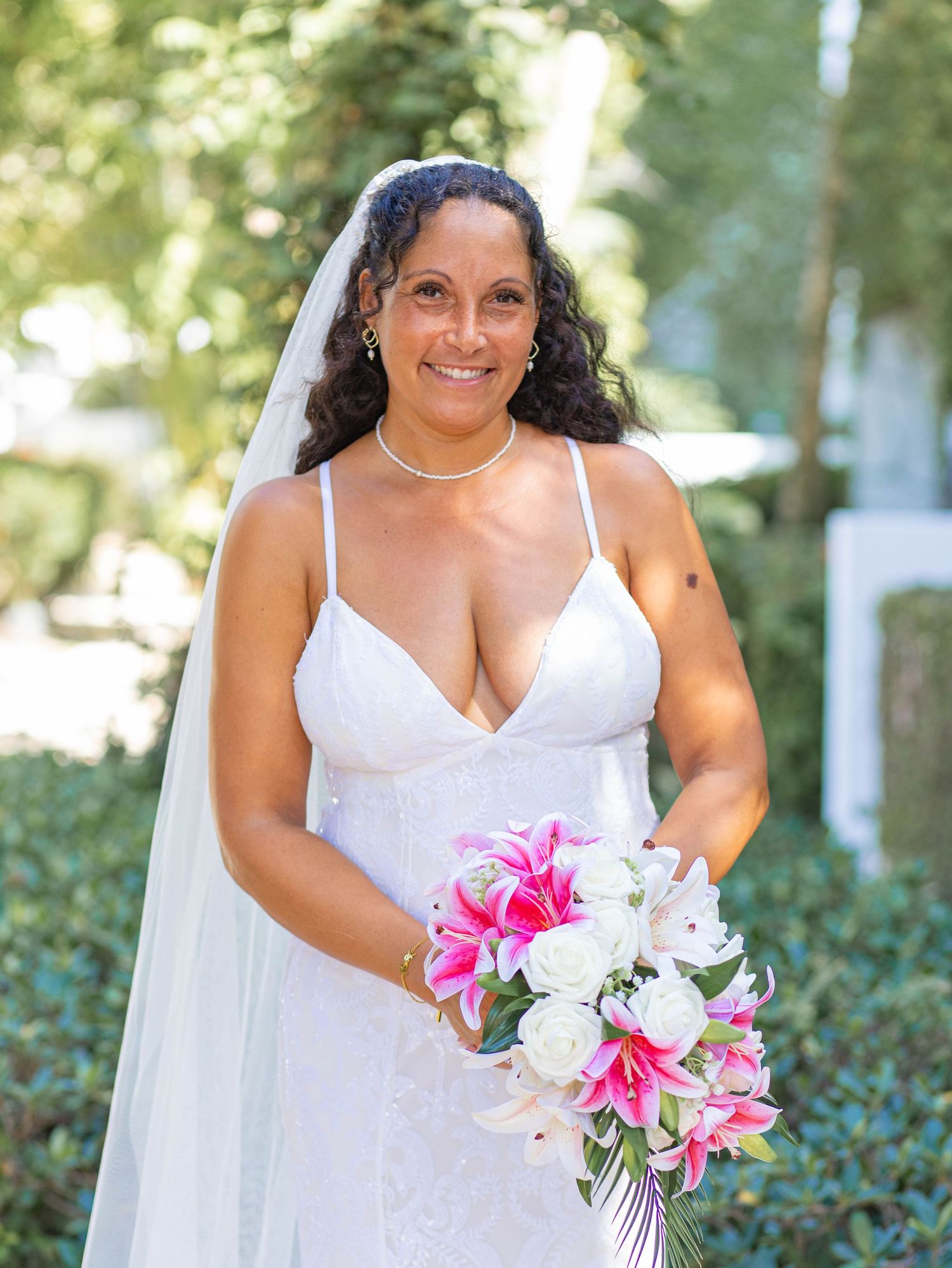 A bride in a white spaghetti strap wedding dress holds a vibrant bouquet of pink and white lilies against a garden backdrop.