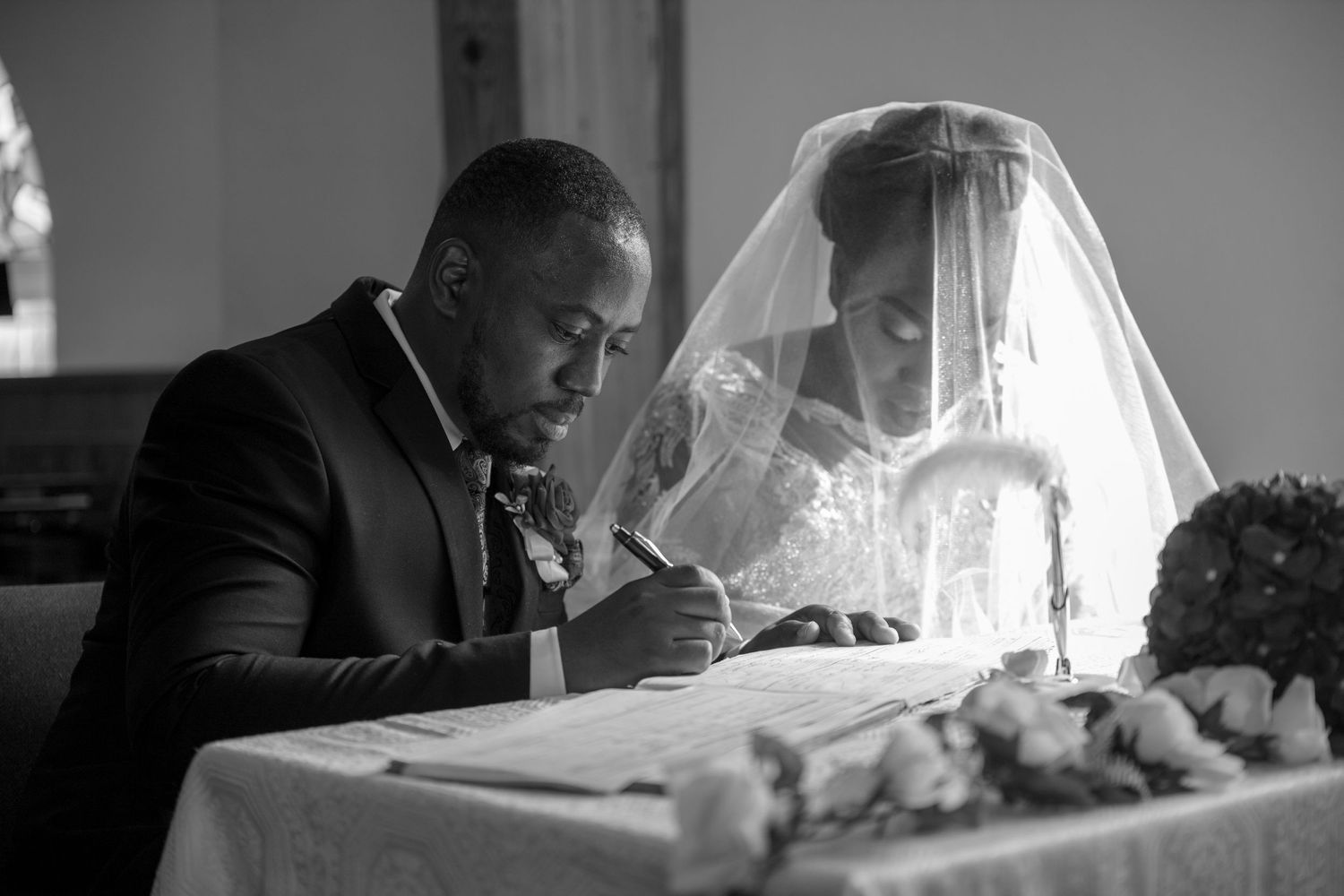 A black and white photograph of a wedding ceremony showing a couple signing documents at a table.