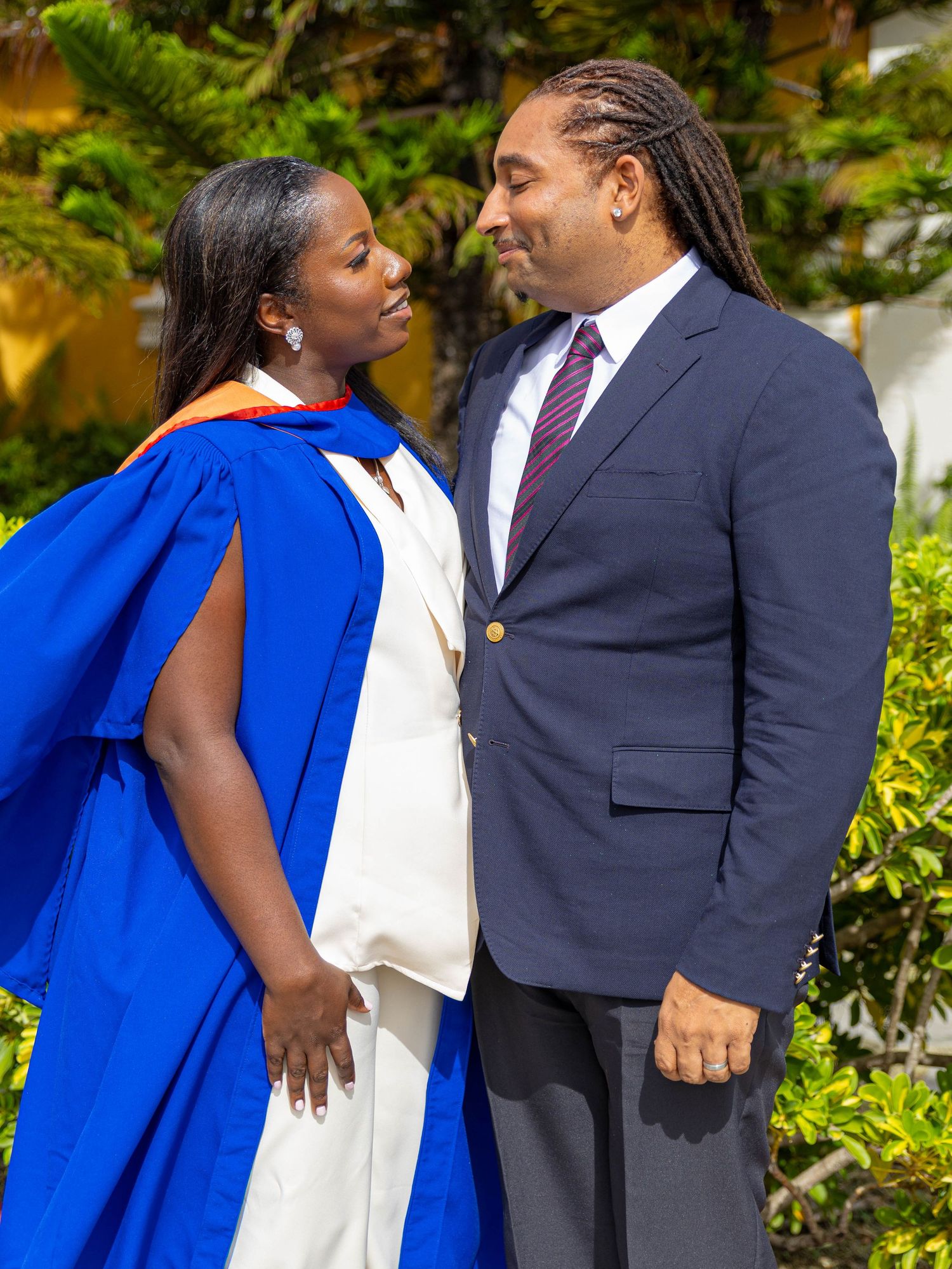 Graduate in blue academic regalia smiles at suited man during outdoor graduation celebration ceremony.