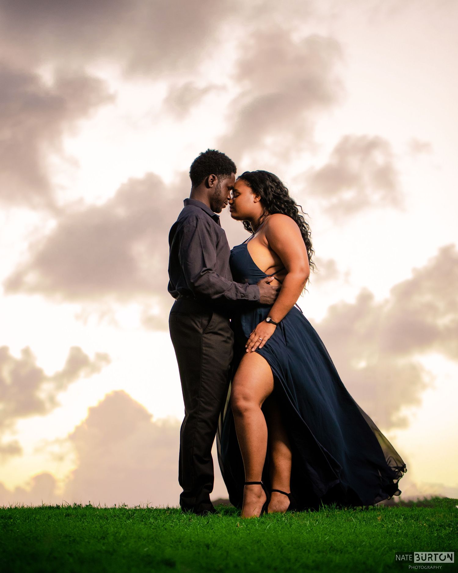 Romantic couple embraces on grassy hilltop during sunset with dramatic cloudy sky behind them.