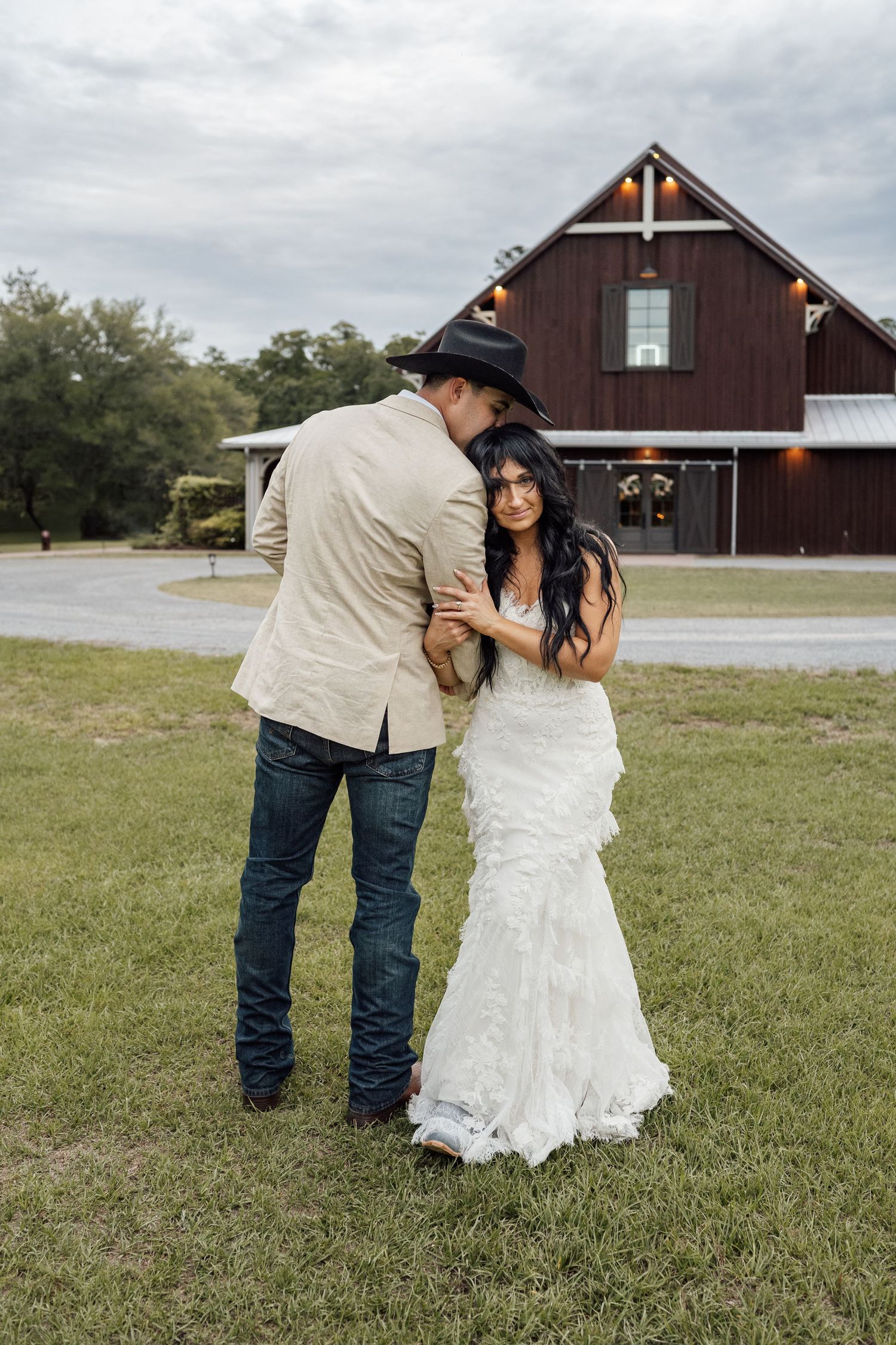 Bride in a ruffled white wedding gown leans into her cowboy-hatted groom on the lawn in front of a rustic red barn venue.