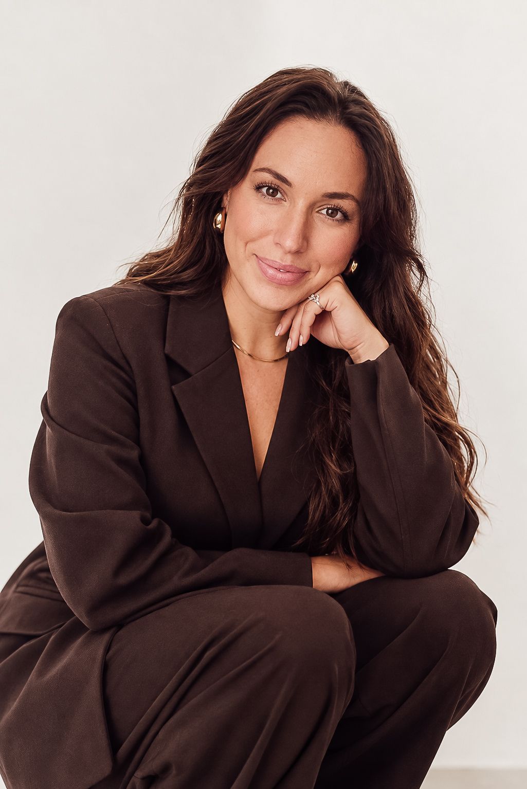 Smiling woman with long wavy brown hair in a brown blazer rests her chin on her hand in a bright studio portrait.