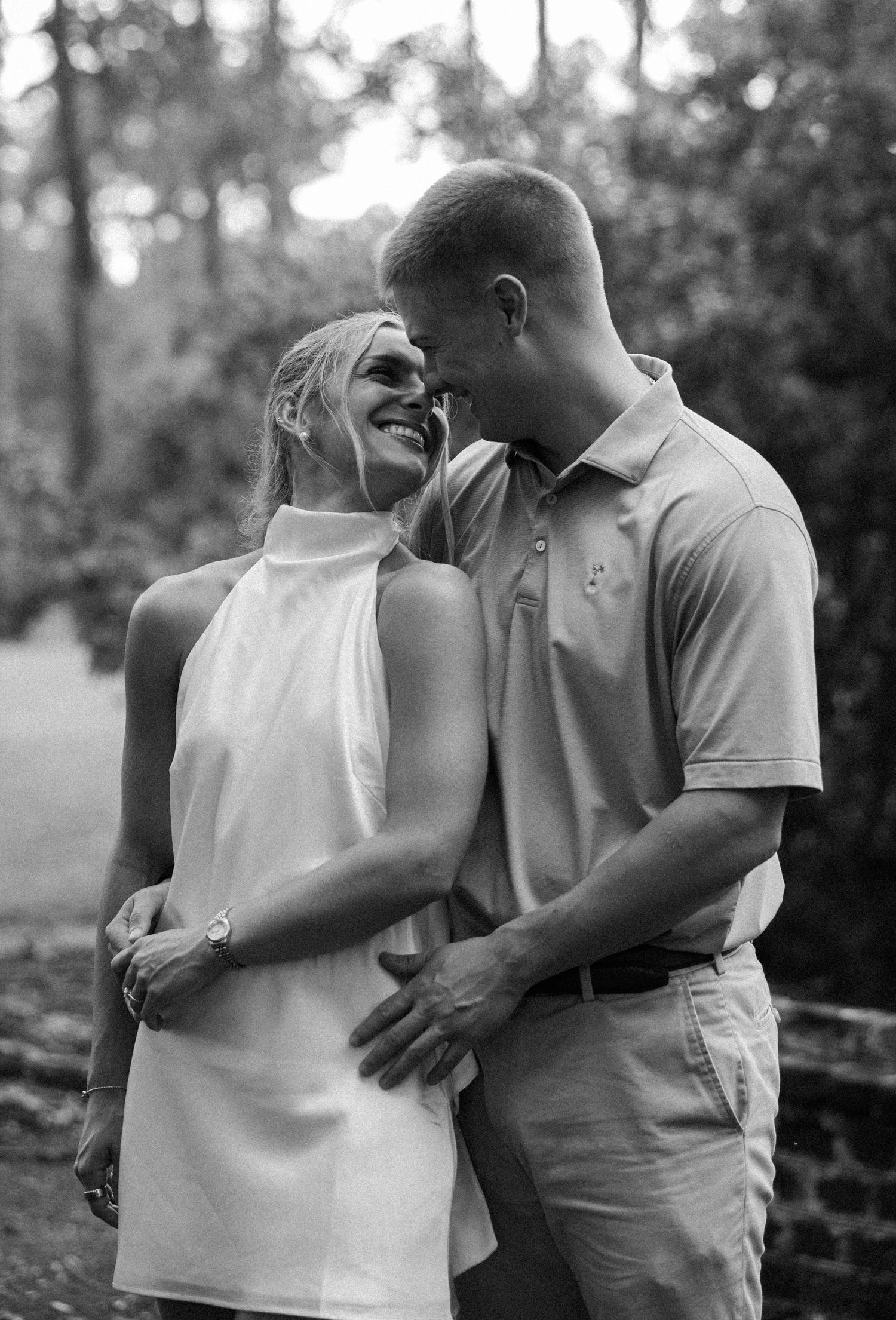 Black and white portrait of a smiling couple embracing closely in a wooded park during an engagement photoshoot.