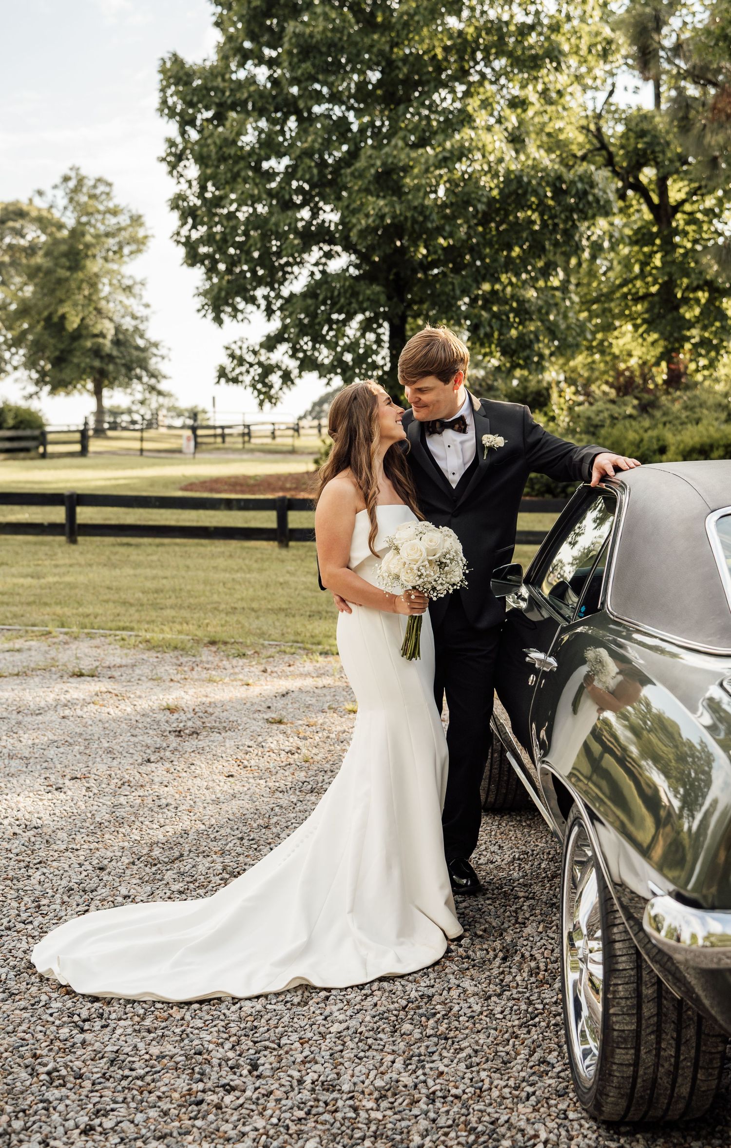 Bride in a fitted white gown with a white bouquet gazes at the groom in a tuxedo beside a vintage car on a gravel drive.