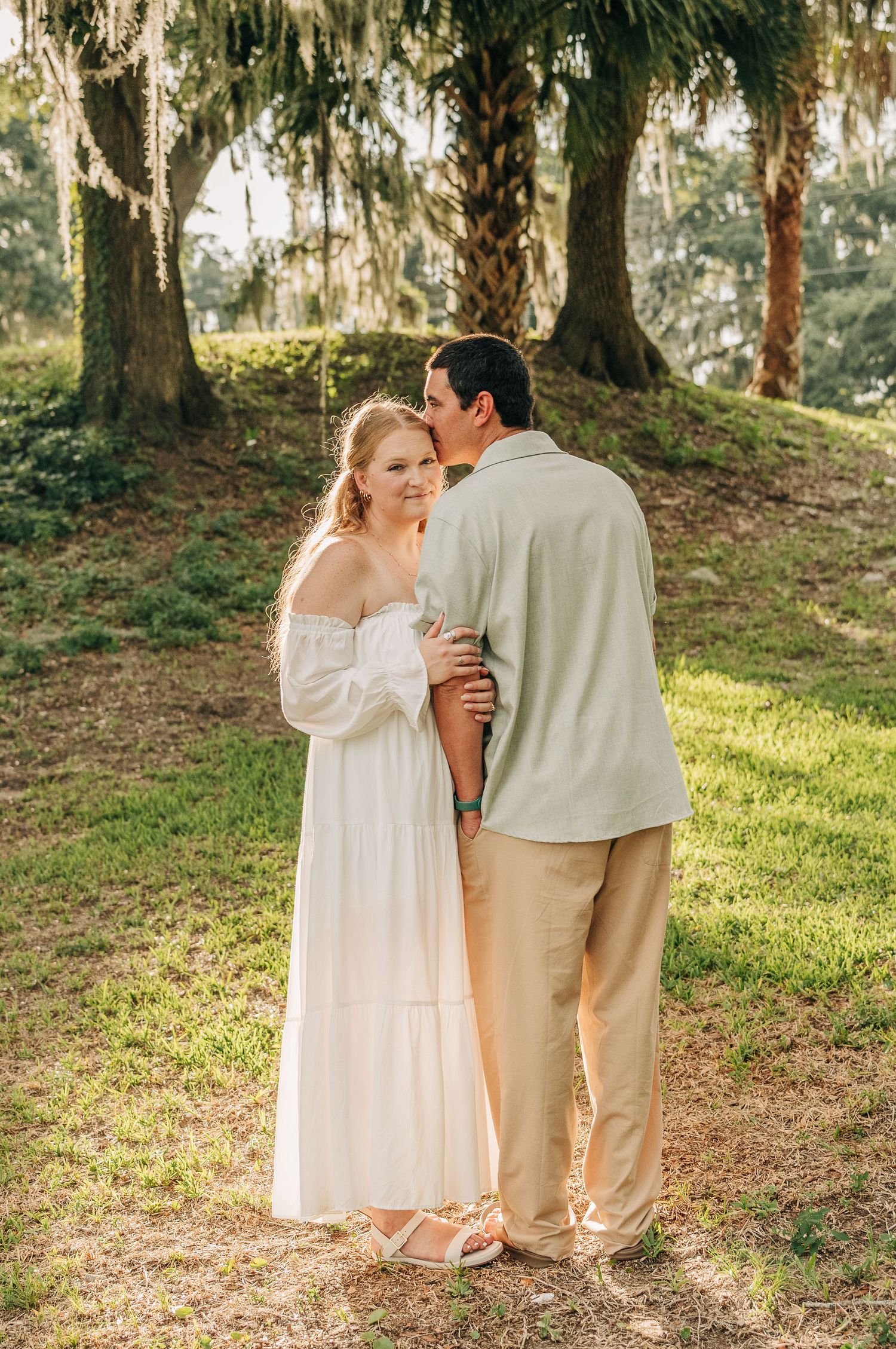 Man kissing his fiancée's forehead in a sunlit grove of palm and oak trees, she wears a white off-shoulder dress.