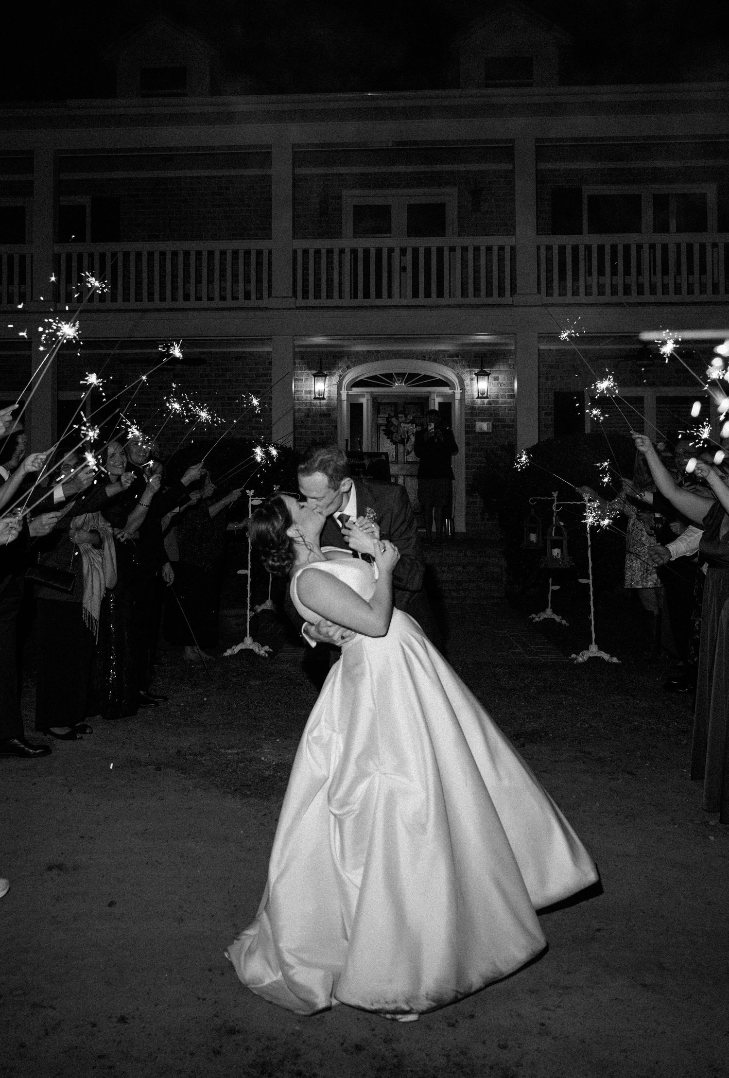 A bride and groom share a kiss in black and white as wedding guests hold sparklers in front of a historic building at night.