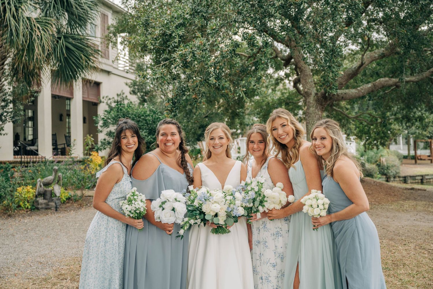 Bride stands among five smiling bridesmaids in dusty blue dresses holding white floral bouquets beneath large oak trees.
