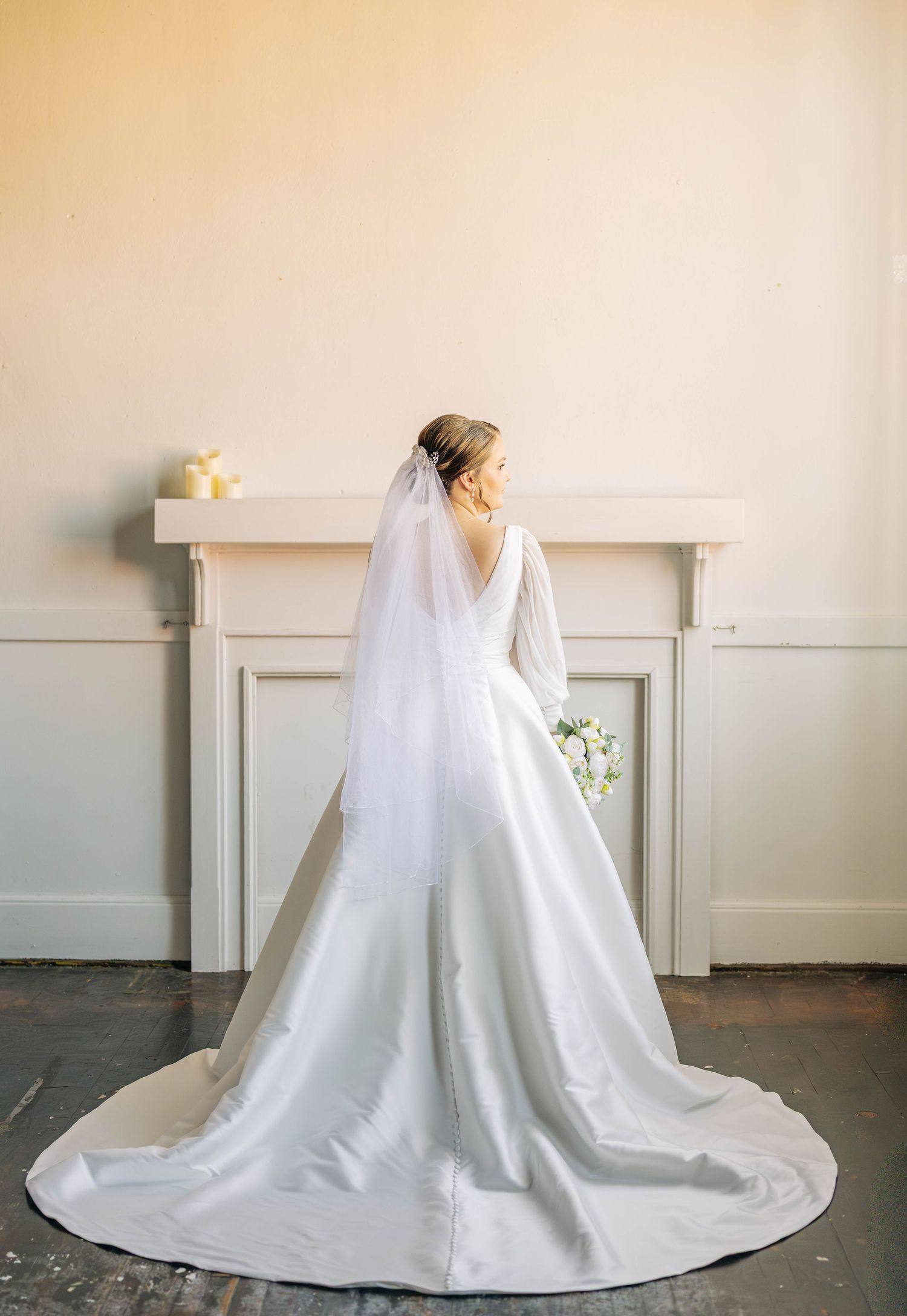 Bride seen from behind in a satin ball gown with a long train and sheer veil, holding a white bouquet beside a mantel.